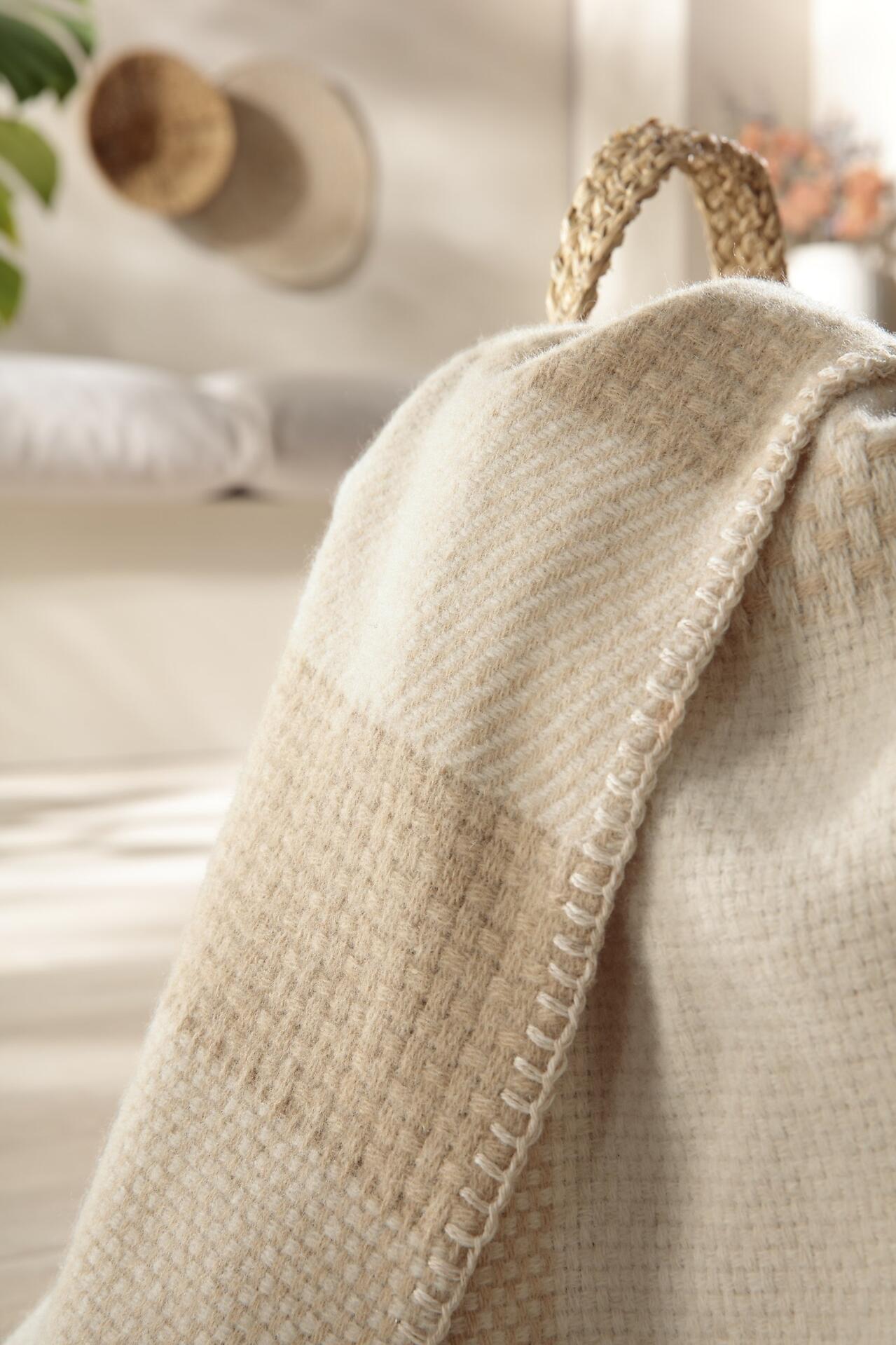 Beige Plaid aus 100% Wolle von Biederlack, detailreich mit grob gewebter Struktur und fransiger Paspelierung über einem Korb drapiert.