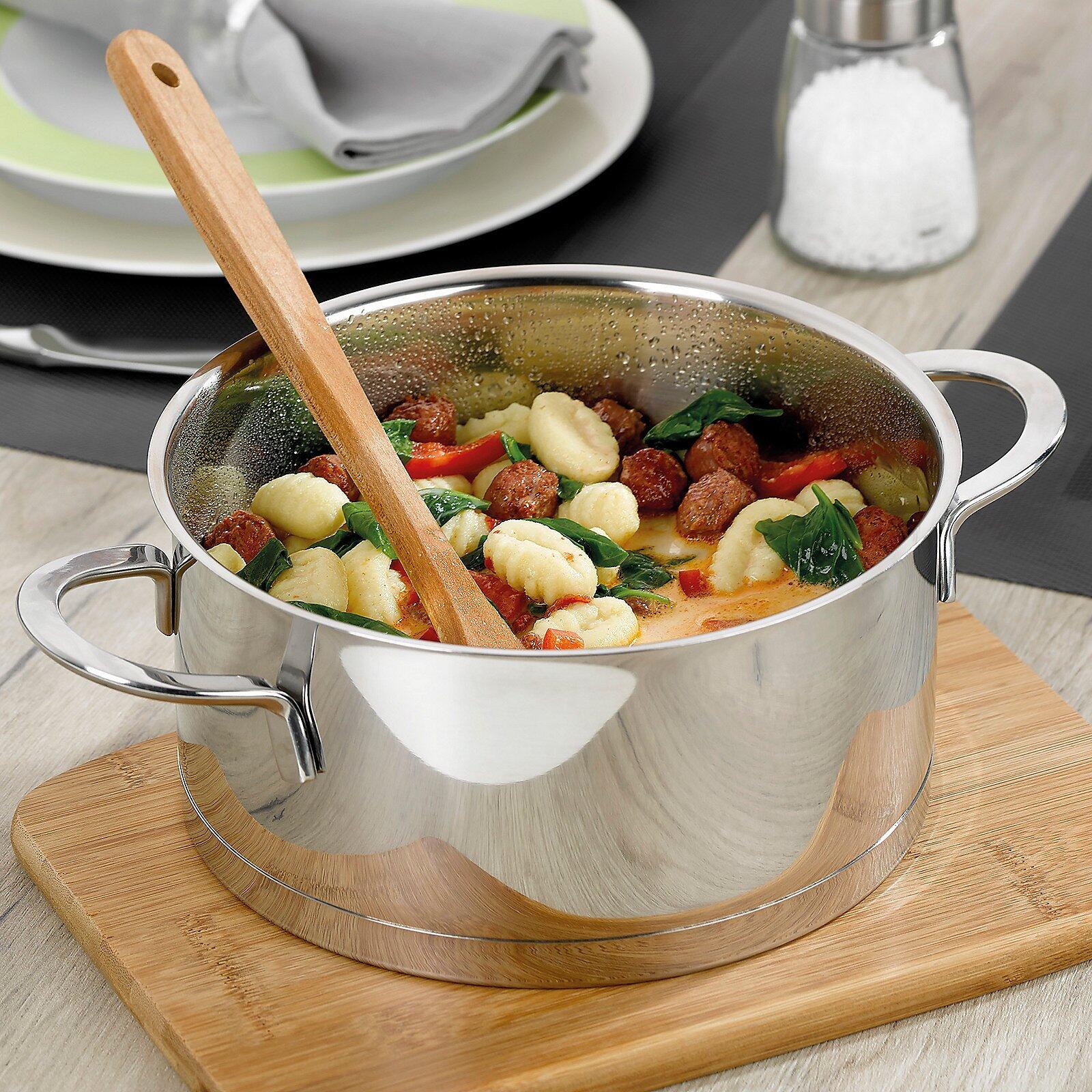 Fleischtopf Cailin aus Edelstahl mit gebogenem Griff und transparentem Deckel, gefüllt mit Gnocchi, Spinat und Wurst, dargestellt aus leicht unterer Perspektive auf einem Holzbrett.
