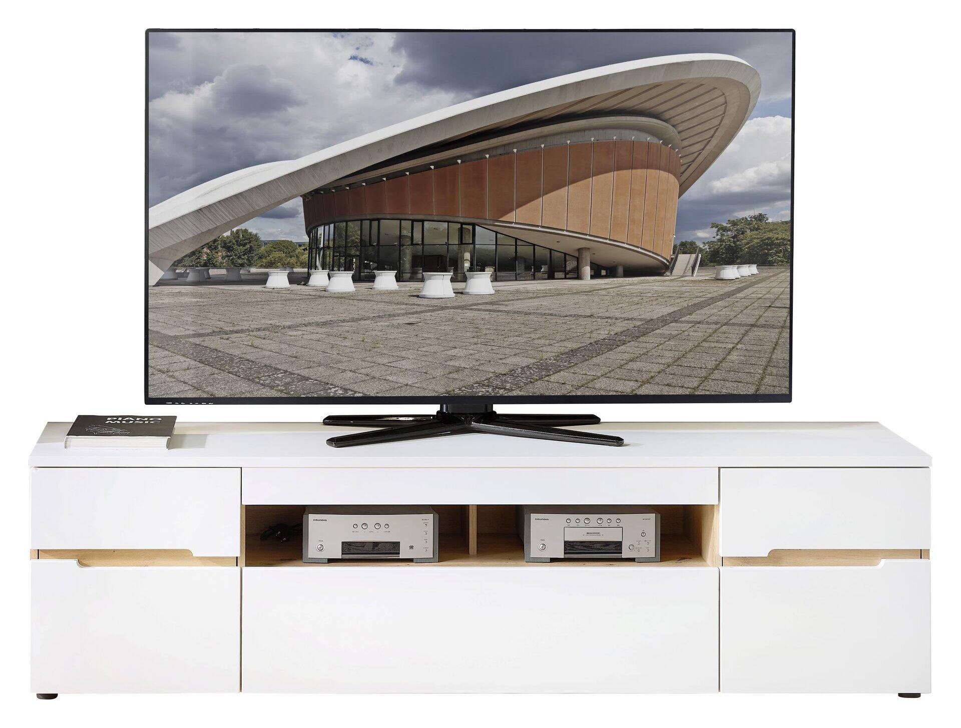 TV-Unterteil Memphis mit weißen Fronten, zwei Holztüren, zwei offenen Fächern und einem Schubkasten, frontal dargestellt mit Blick auf einen Fernseher und Audio-Geräte.