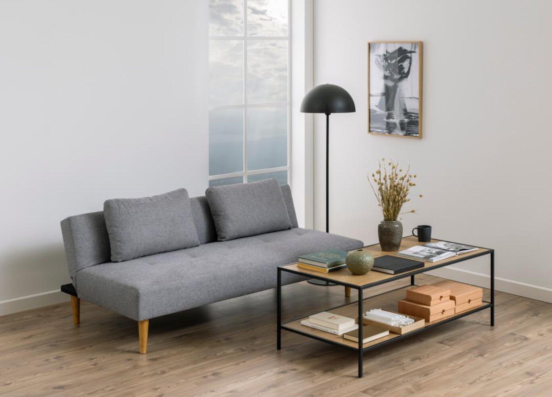 Schlafcouch Lucca in Grau mit schlichter, moderner Optik und leicht erhöhter Rückenlehne, dargestellt in einer seitlichen Perspektive mit angrenzendem Couchtisch und dekorativen Elementen.