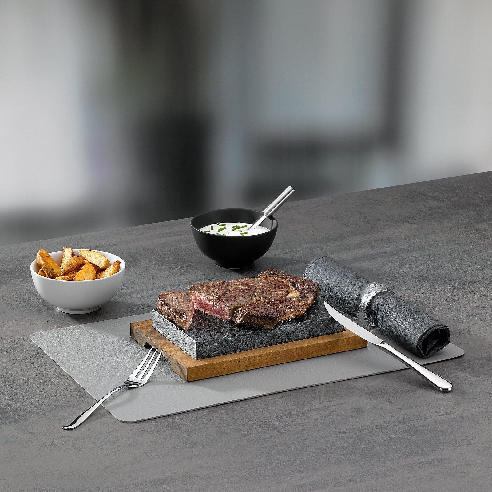 Steak auf Holzbrett mit Besteck und Soße, präsentiert auf einer grauen Unterlage.