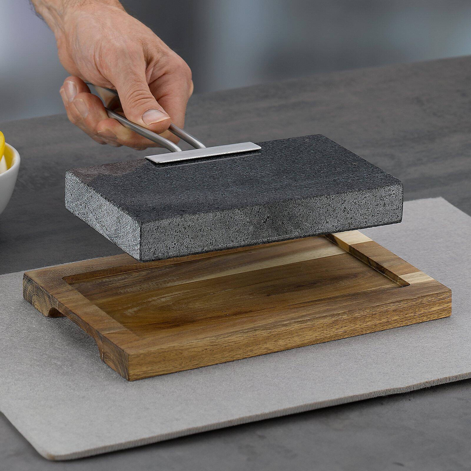 Quadratische schwarze Steinplatte auf Holzbrettablage mit integrierten Vertiefungen, gehalten von einer Hand.