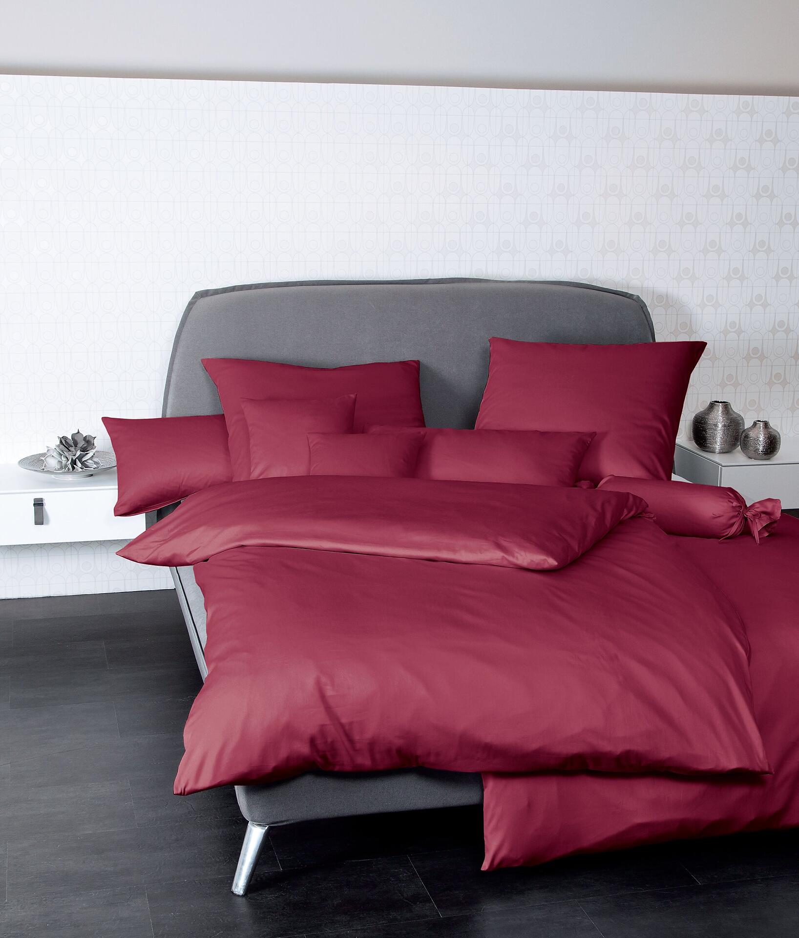 Bordeauxfarbenes Kissen "Colors" aus Mako-Satin-Baumwolle, im ¾-Ansicht auf einem stilvollen Bett gestapelt, um die luxuriöse Bettwäsche und den entspannten Look zu präsentieren.