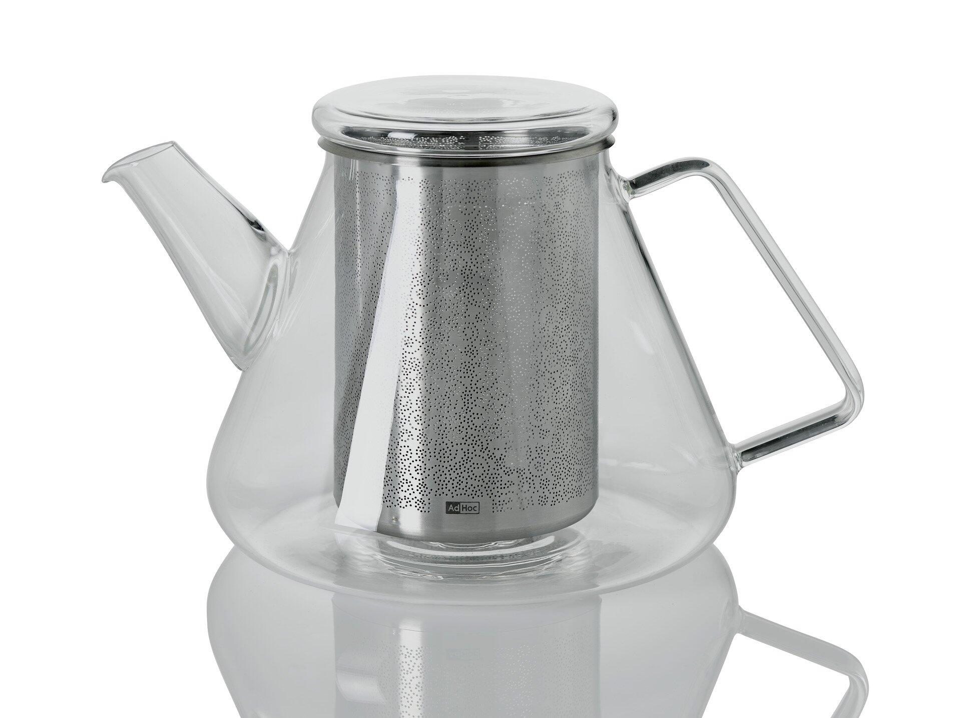 Teekessel Orient+ aus Glas und Edelstahl mit silberfarbenem Griff und Deckel, dessen Löcher sichtbar sind, auf einer reflektierenden Oberfläche dargestellt.