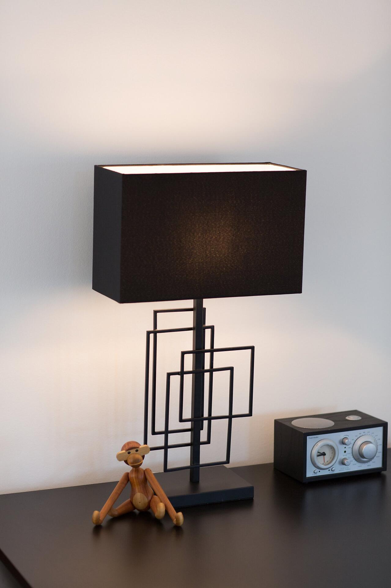 Tischleuchte Paragon mit modernem, geometrischem Design und filigranem Metallgestell, die vor einem weißen Hintergrund auf einem Nachttisch steht und weiche Schatten wirft.