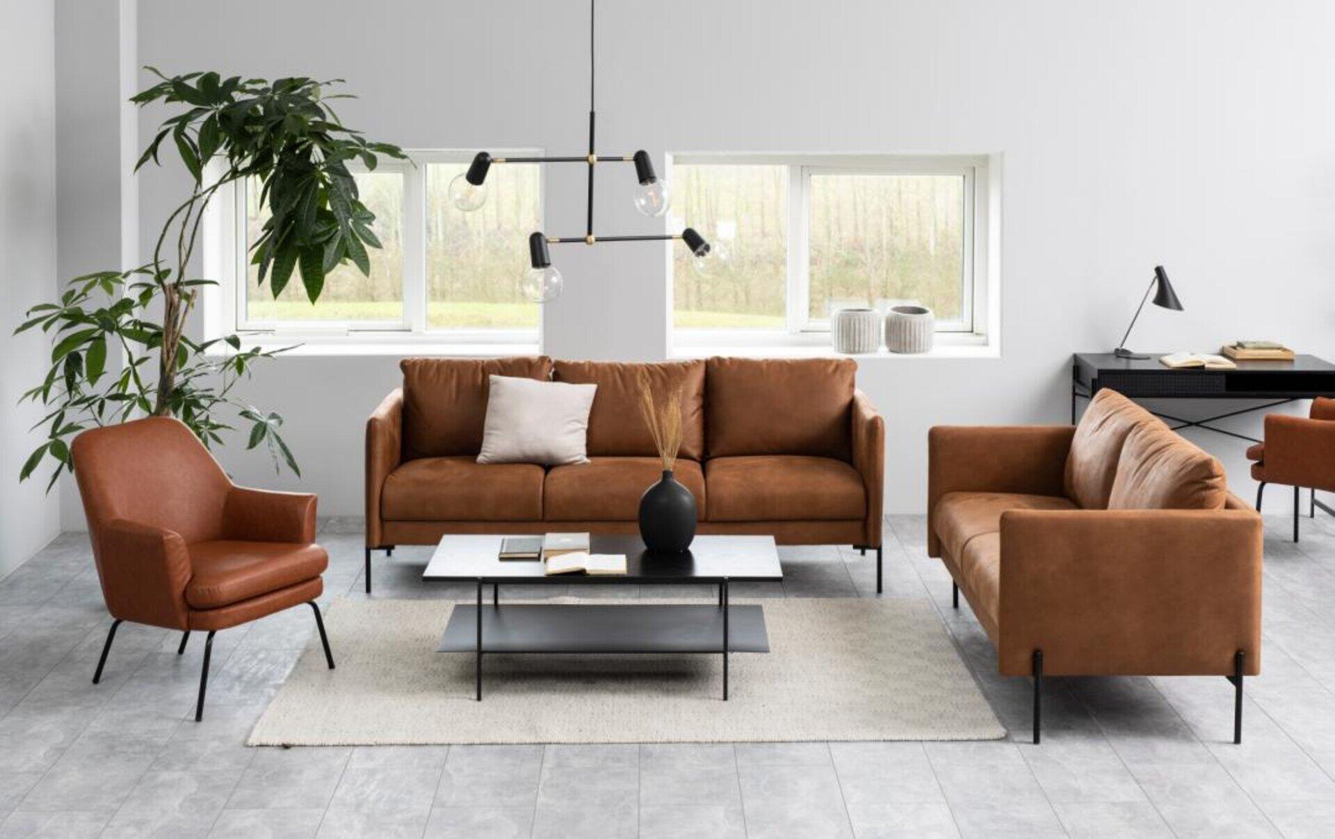 Couchtisch Angus mit schwarzer Tischplatte und metallischen Beinen, im Raum arrangiert mit Sofa, Sessel und Pflanze.