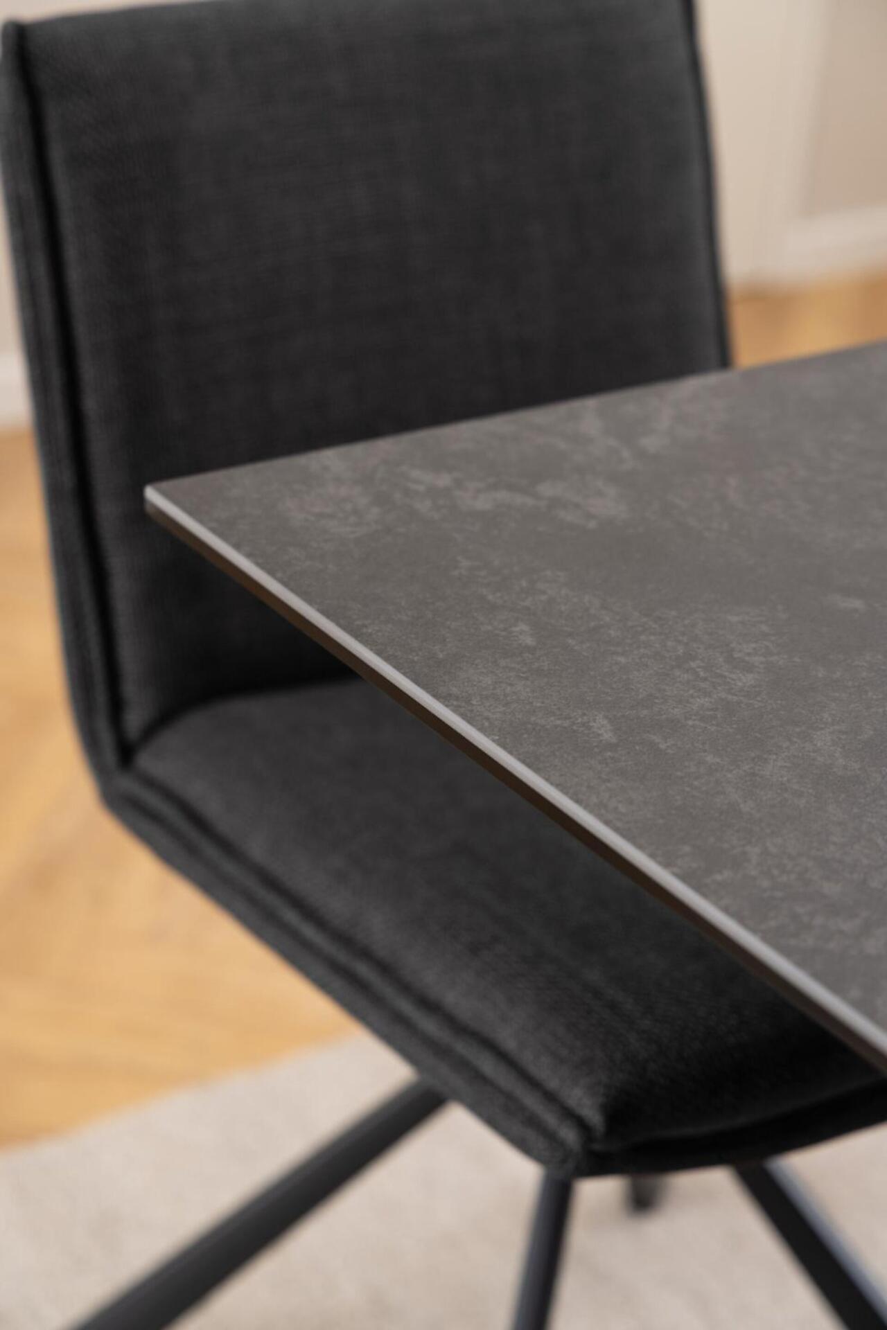 Detailaufnahme des Esstisches Heaven: Dunkle, strukturierte Tischplatte in Kombination mit einem dunkelgrauen, samtigen Stuhl mit schwarzem Metallrahmen.