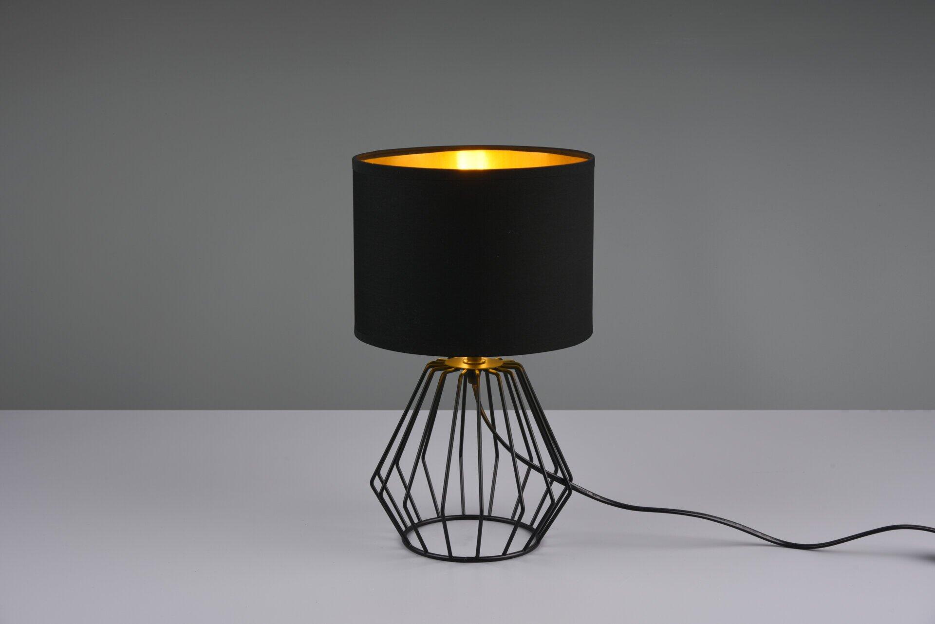 Tischleuchte Chuck mit zylindrischem Lampenschirm aus dunklem Stoff und geometrischem Drahtgestell auf goldener Plattform.