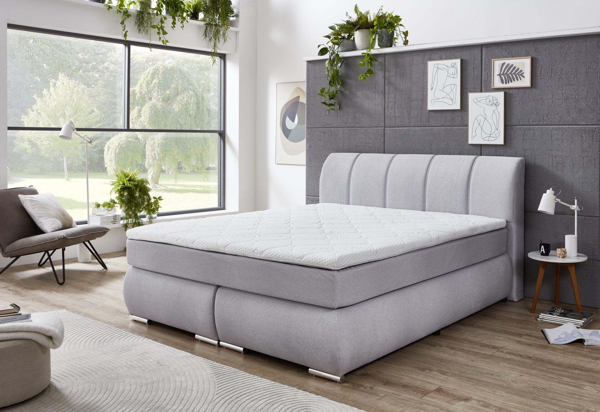 Hellgraues Boxspringbett Graz BX2630 mit hochgewölbtem, gepolstertem Kopfteil und seitlichen Mechanismen für integrierten Stauraum, im Schlafzimmerambiente.
