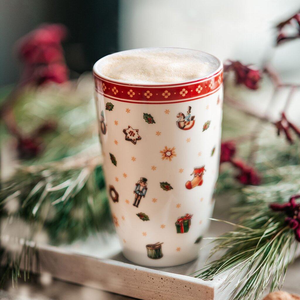 Becher Toys Delight mit festlichem Dekor aus Nussknackern, Äpfeln und Sternen auf hellgelbem Hintergrund, gefüllt mit Kaffee, auf einem Tablett mit Tannenzweigen.