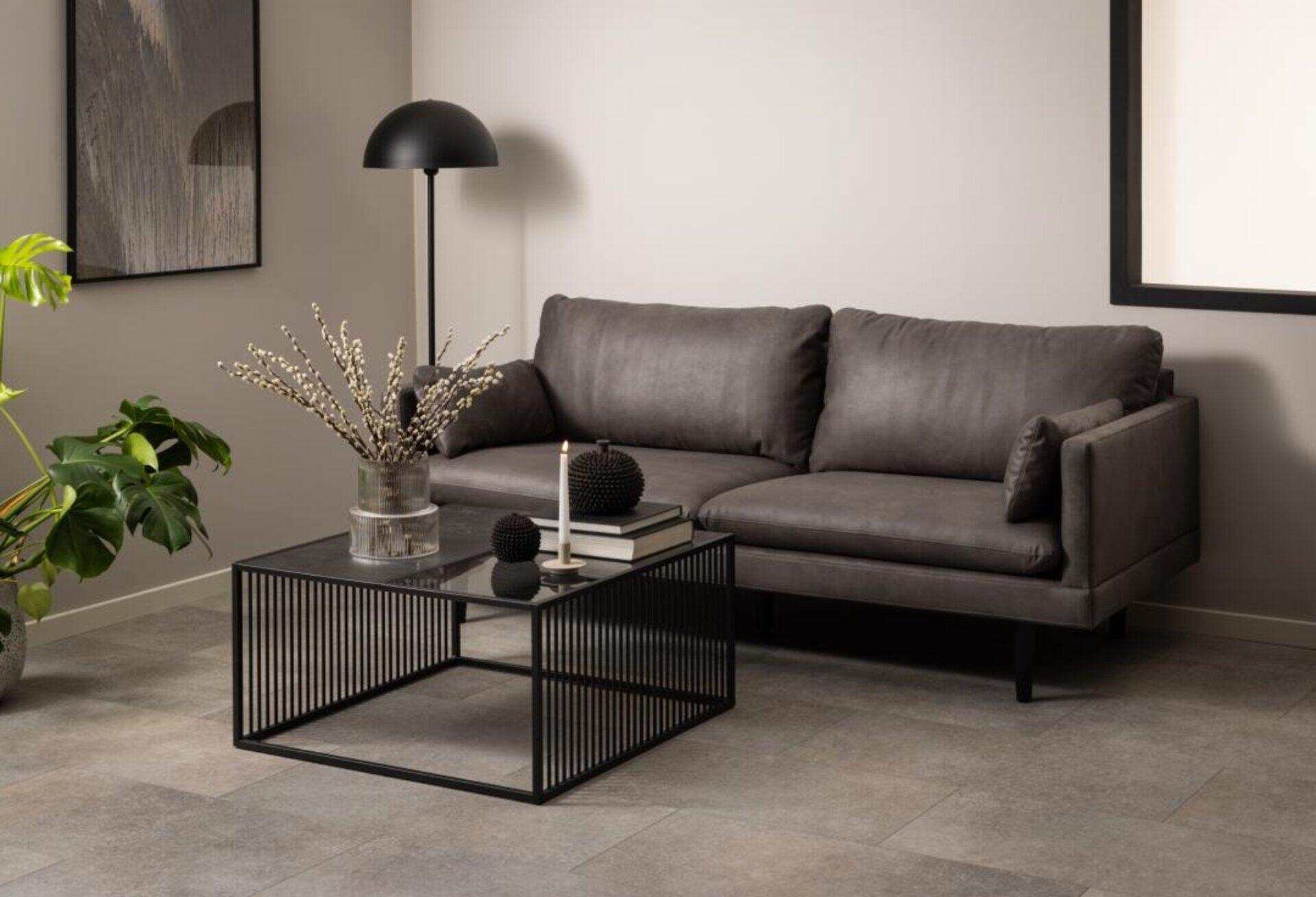 Couchtisch Strington mit schwarzem Metallgestell und Glasplatte vor einer grauen Eckcouch in einem modernen Wohnzimmer.
