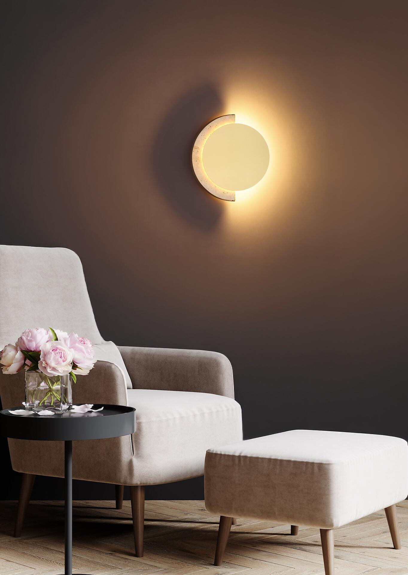 Runde Wandleuchte ROJAS mit Travertin-Glas und warmweißem LED-Licht, dargestellt an einer Wand vor einem Sessel und Beistelltisch.