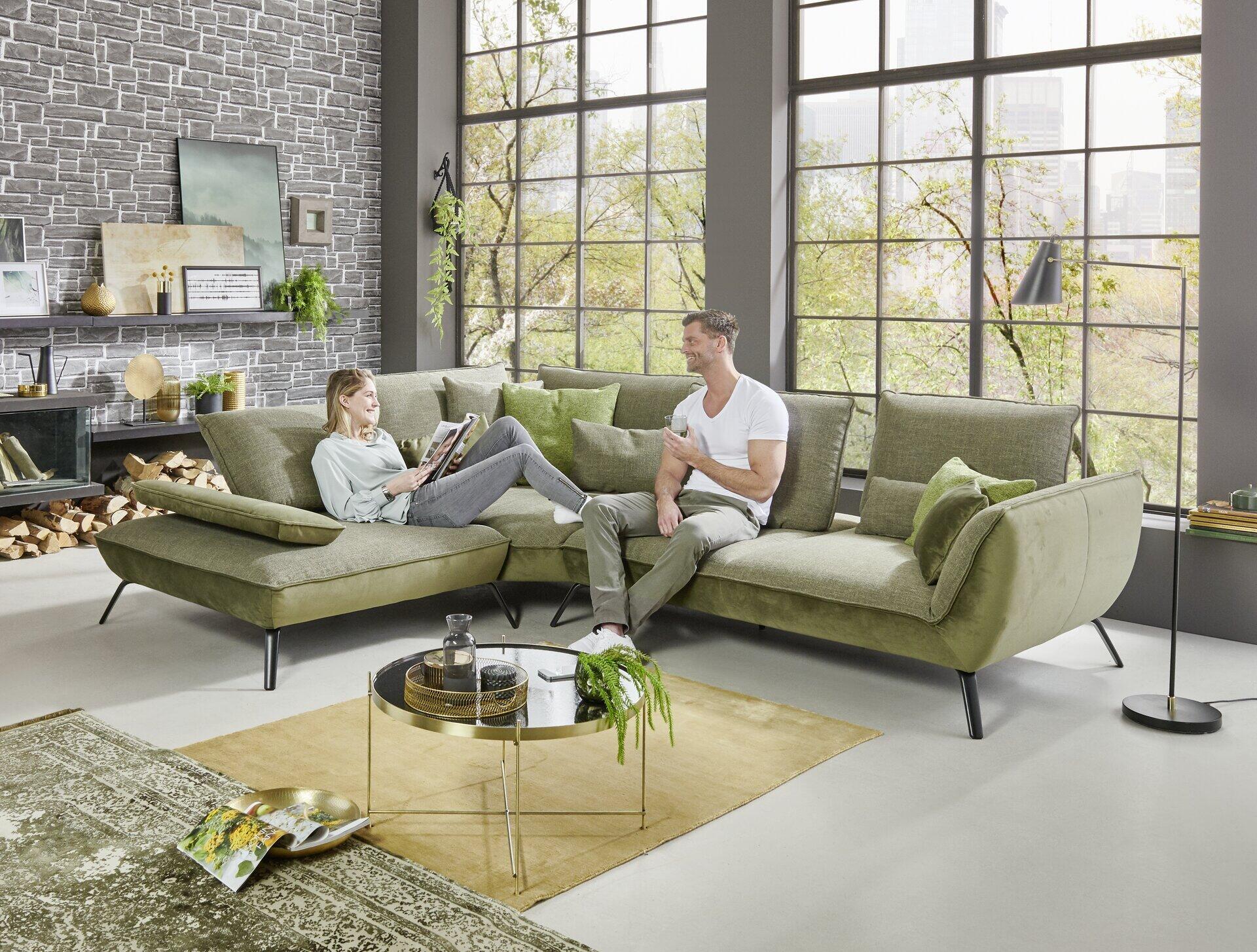 Moderne Polstergarnitur Alpha Plus in Olivgrün mit schwarzen Metallfüßen und flexiblen Elementen für verschiedene Sitzmöglichkeiten, dargestellt in einer frontal leicht erhöhten Perspektive mit zwei Personen auf der Couch.