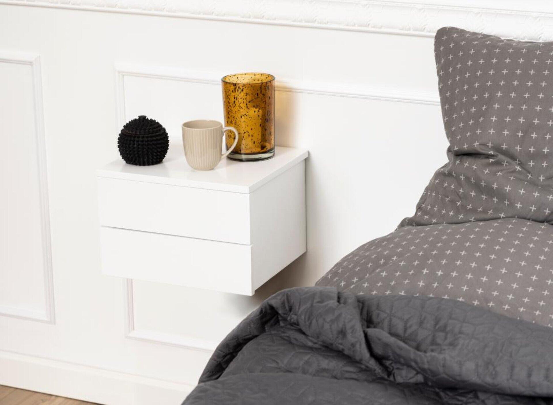 Nachttisch Avignon in Weiß, schwebend an der Wand mit einer Schublade, ergänzt durch eine Tasse, Kerze und Dekofigur.