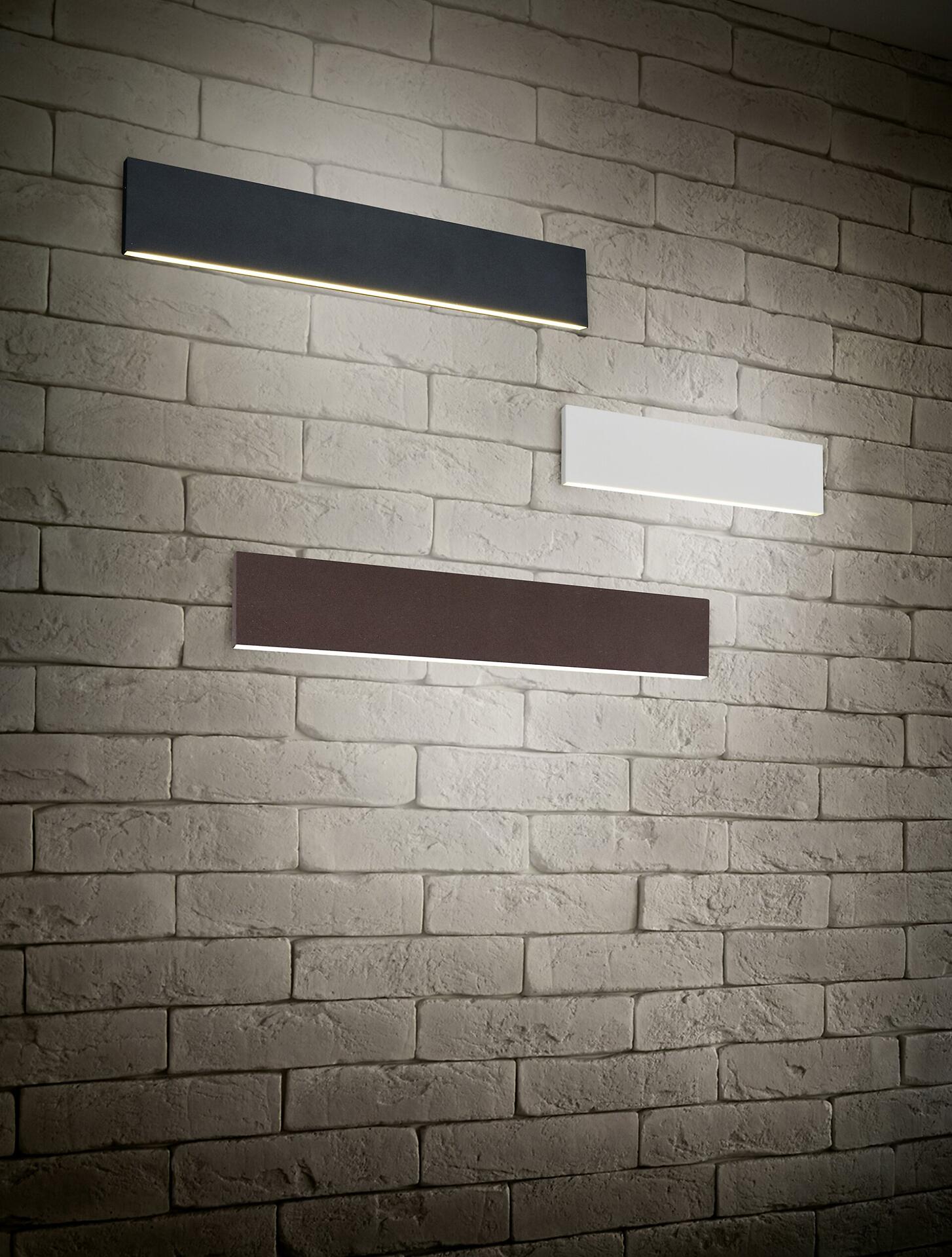 Zwei Wandleuchten Concha mit rechteckigem Design und indirekter Beleuchtung, montiert an einer hellgrauen Ziegelwand, wobei der Blickwinkel leicht von unten die schlanken Leuchten hervorhebt.