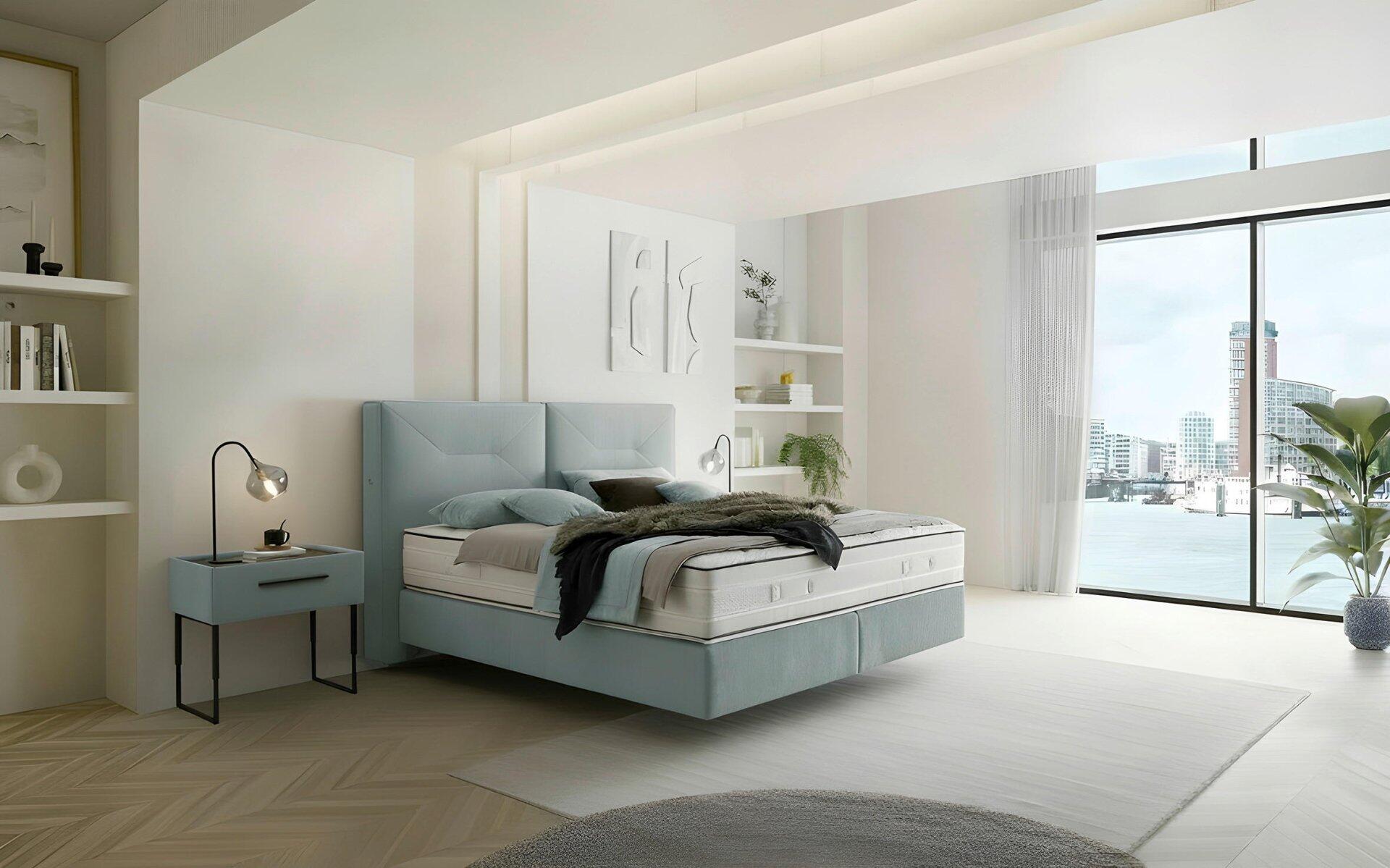 Luxor Line Boxspringbett mit hellblau gestepptem Kopfteil und Schwebeoptik-Fuß, integriert in ein modernes Schlafzimmer mit Hafenblick.