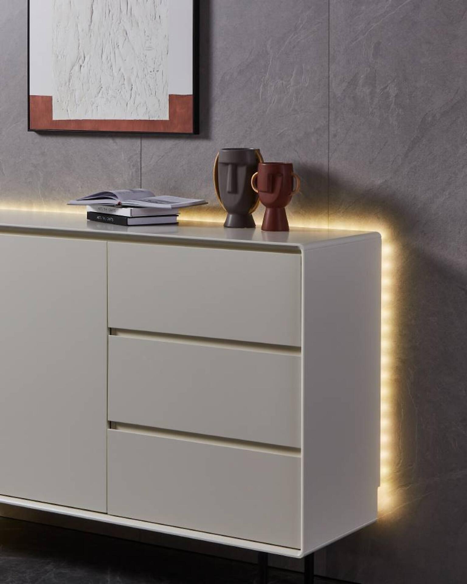 Kommode Stripe, Led Stripe-Z mit cremefarbenem Korpus, schwarzen Metallfüßen und integrierter LED-Leiste, die ein warmes Licht an die Wand wirft.