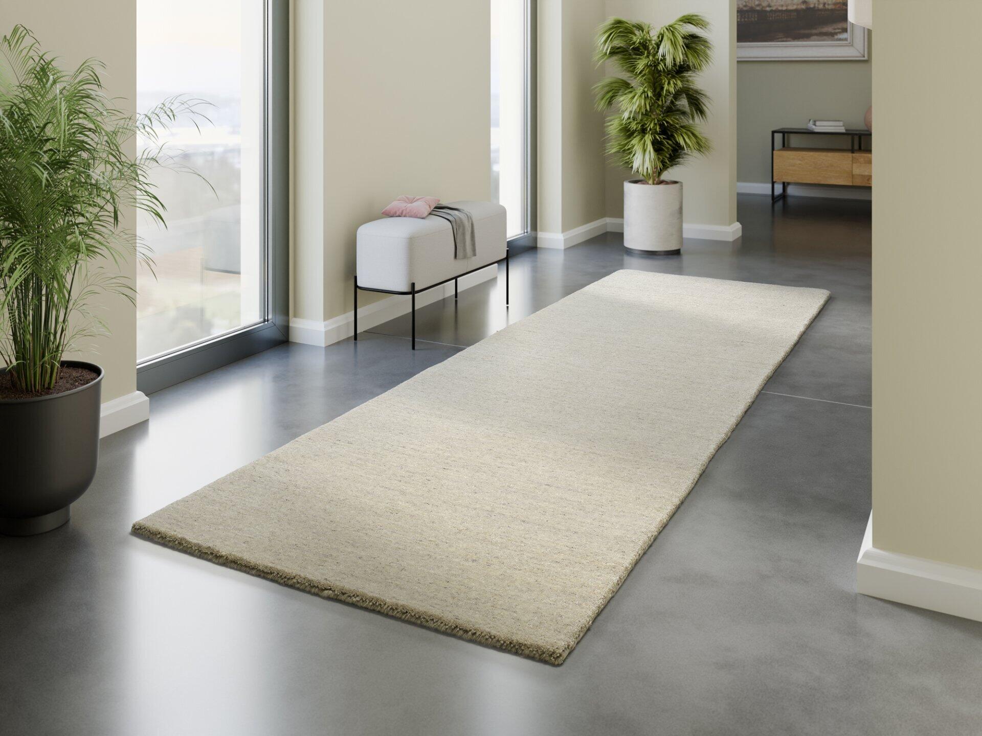 Handloom Ararat Wollteppich in Beige-Grau, diagonal auf einem Steinboden liegend, mit Pflanze und Hocker im Hintergrund.