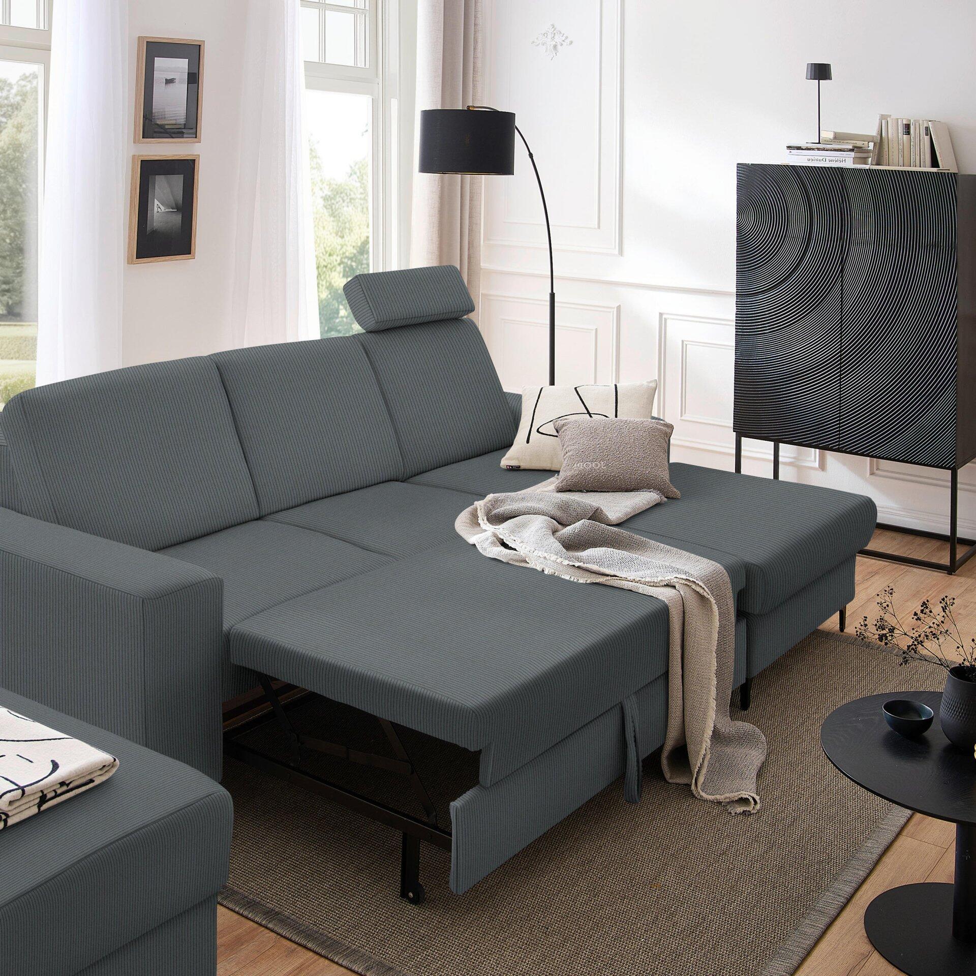 Ecksofa Balina in Grau mit ausgeklappter Schlaffunktion und Winkelfüßen, leicht seitliche Ansicht mit Teppich und Sideboard im Hintergrund.