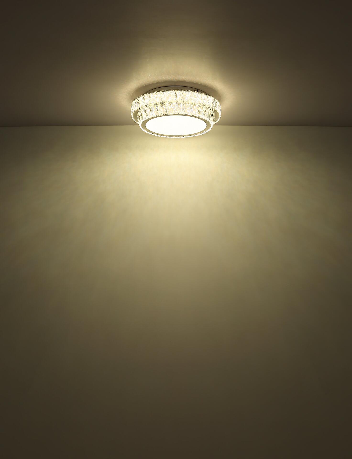 Deckenleuchte KLARA mit weißem Korpus und glitzerndem Kristallring, die in Frontalperspektive warmweißes Licht und einen hellen Schein an der Wand zeigt.