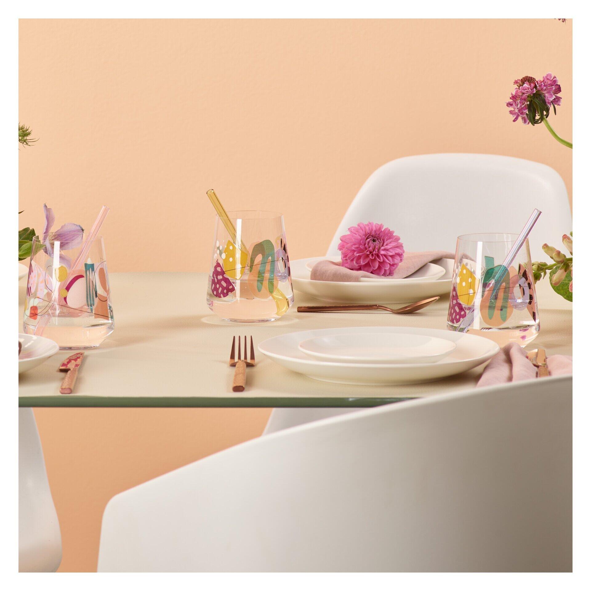 Vier bunte Universalgläser mit gemustertem Design, präsentiert auf einer Tafel mit dekorativen Elementen und einem weißen Stuhl vor hellrosa Hintergrund.