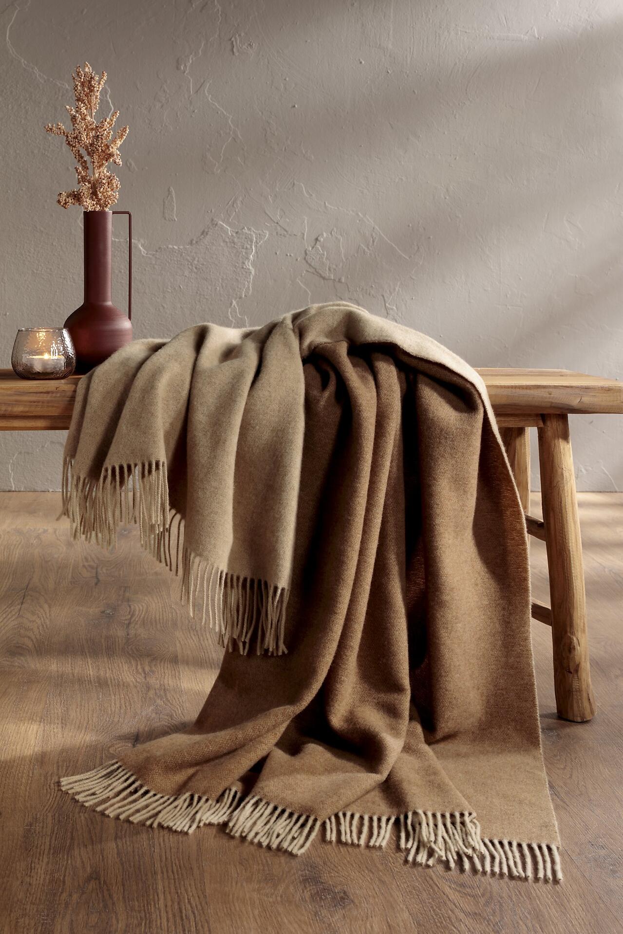Gemütliche camel-beige Plaid-Decke mit Fransen auf einem rustikalen Holzhocker, Detailaufnahme der wolligen Textur.