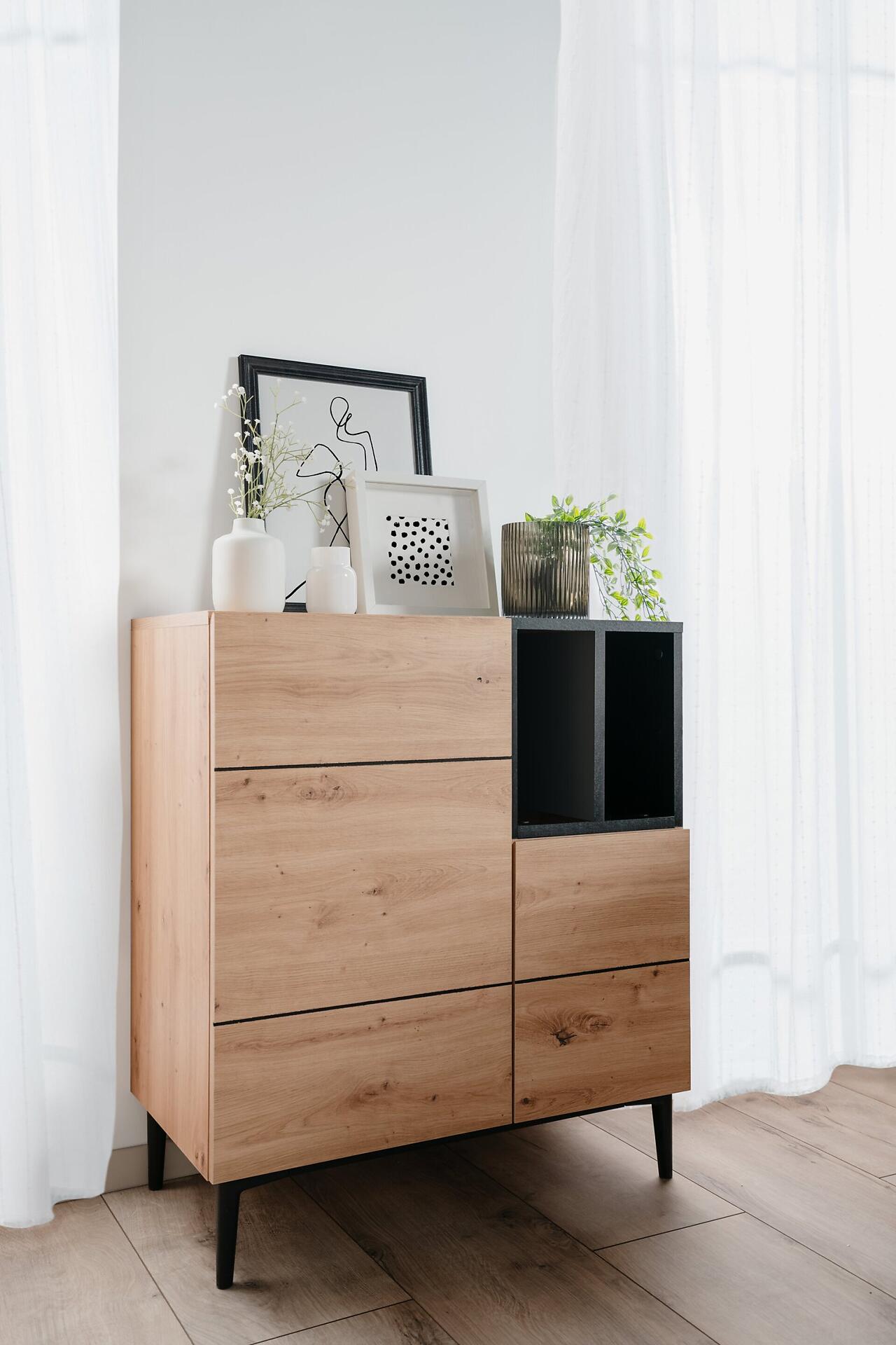 Helle Holzkommode Nola 01A mit schwarzen Füßen, drei Schubladen, offenem Fach und griffarmer Gestaltung, leicht seitlich und von unten aufgenommen.