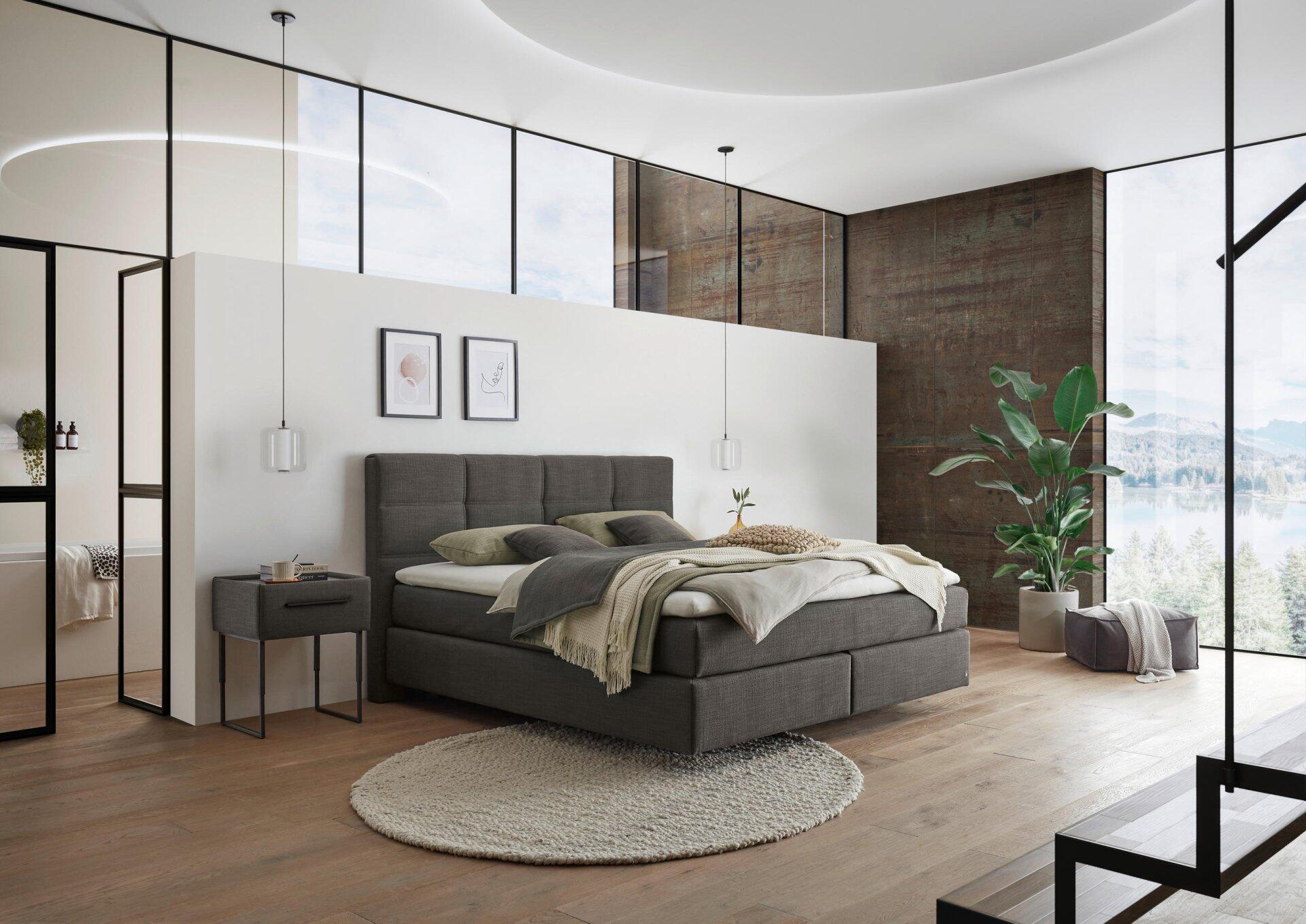 Boxspringbett Luxor Line BX mit grauem Stoffbezug, hohem Kopfteil und Nachttisch am Fußende, aufgenommen aus erhöhter Perspektive im modernen Schlafzimmer.