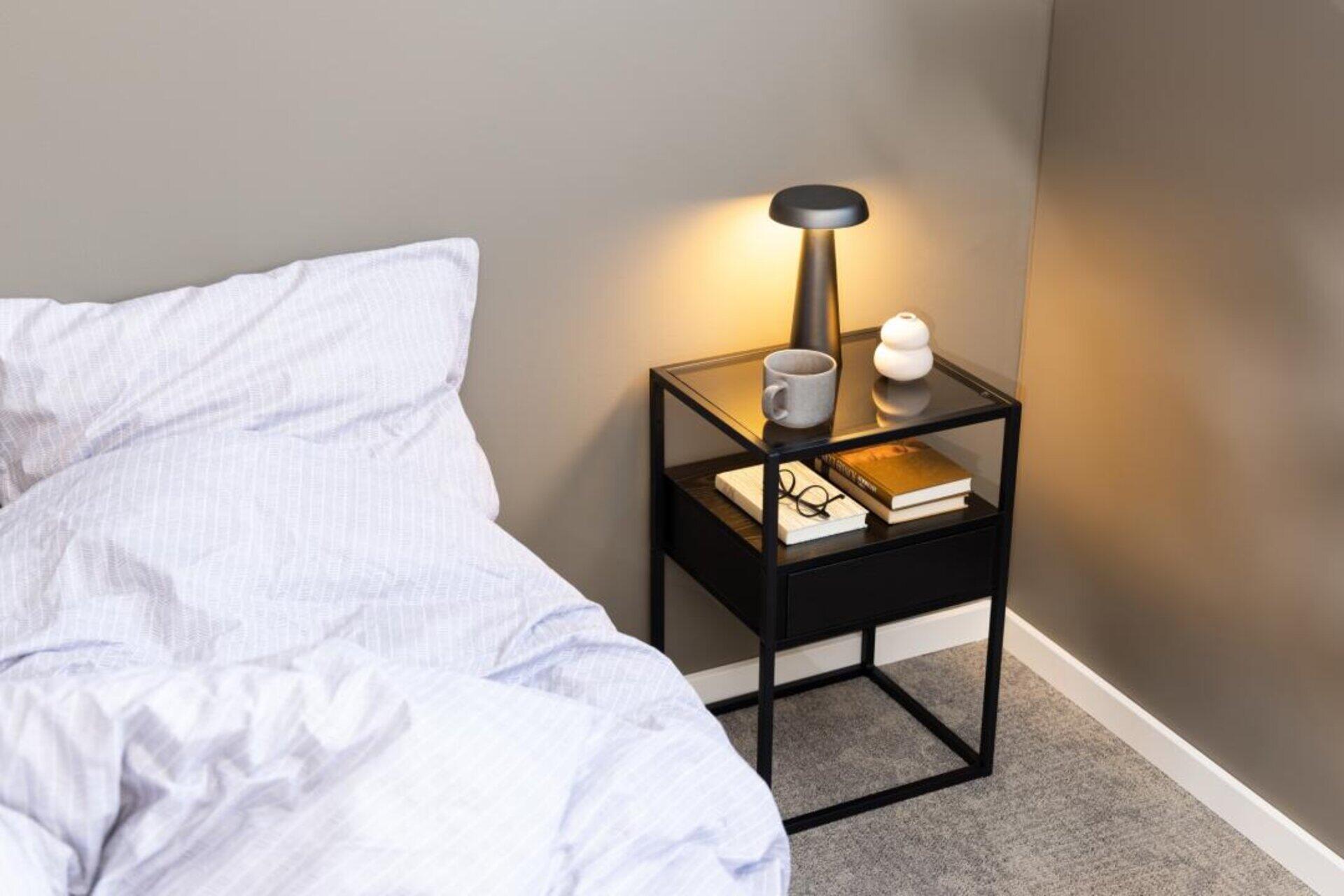 Nachttisch Randolf aus schwarzem Melamin mit einer Schublade und offenem Raum, präsentiert im Schlafzimmer mit Tischlampe und Dekor.