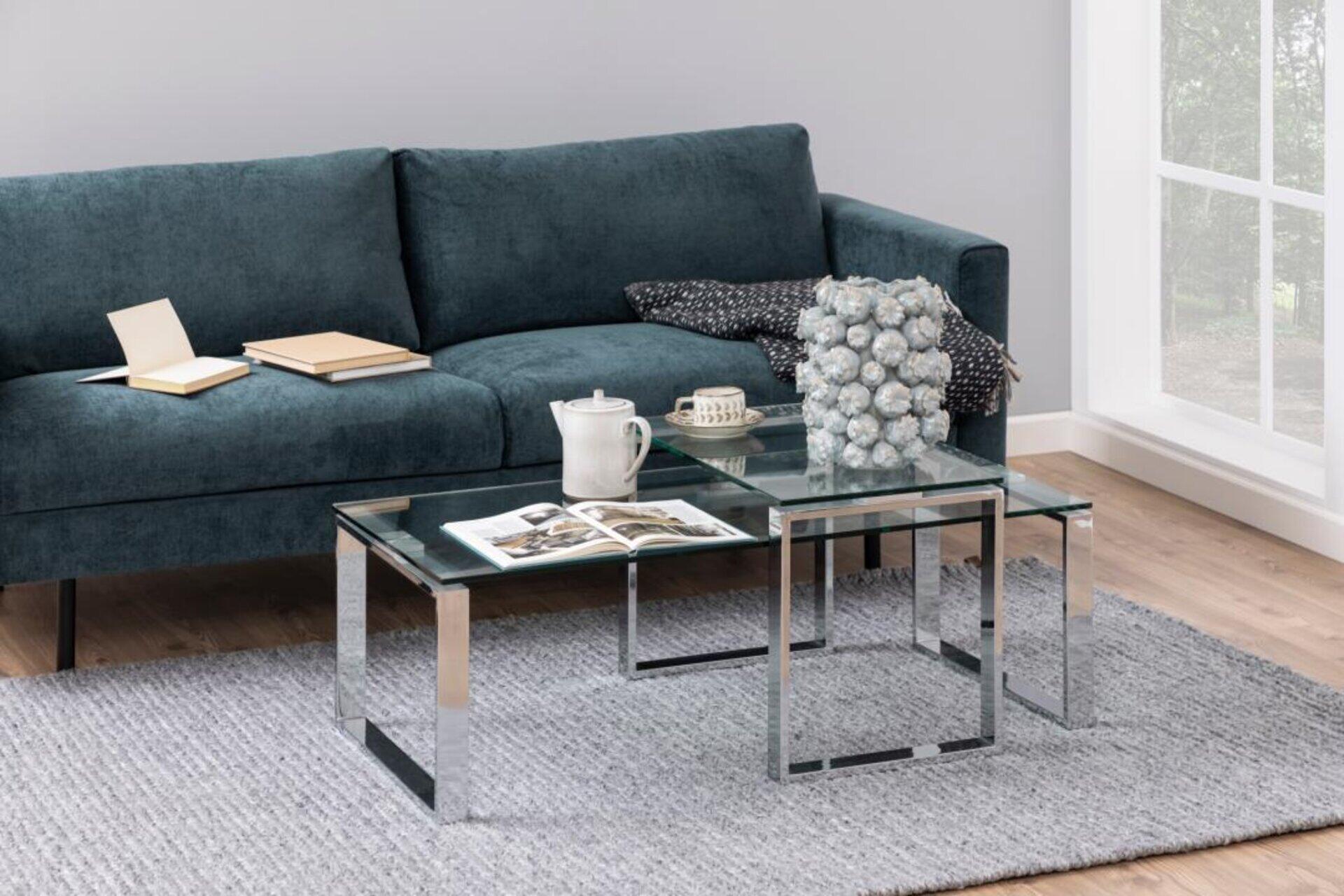 Couchtisch Katrine: Set aus zwei Tischen mit transparenter Glasplatte und silbernem Metallgestell, dargestellt in einem Wohnzimmerbereich aus leicht erhöhter Perspektive.