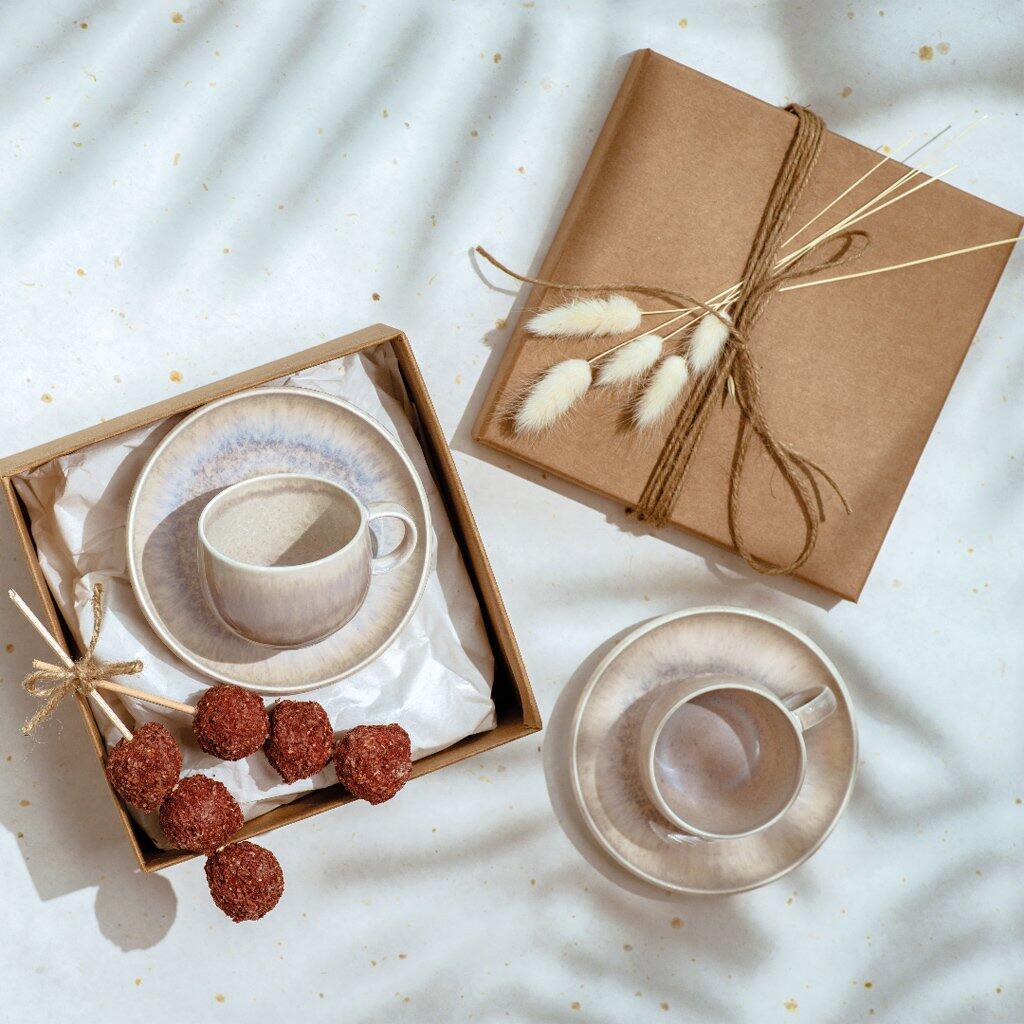 Espressotasse "Perlemor Sand" von Villeroy & Boch in Geschenkbox mit Untertasse, Gräsern und roten Snacks, fotografiert aus der Vogelperspektive.