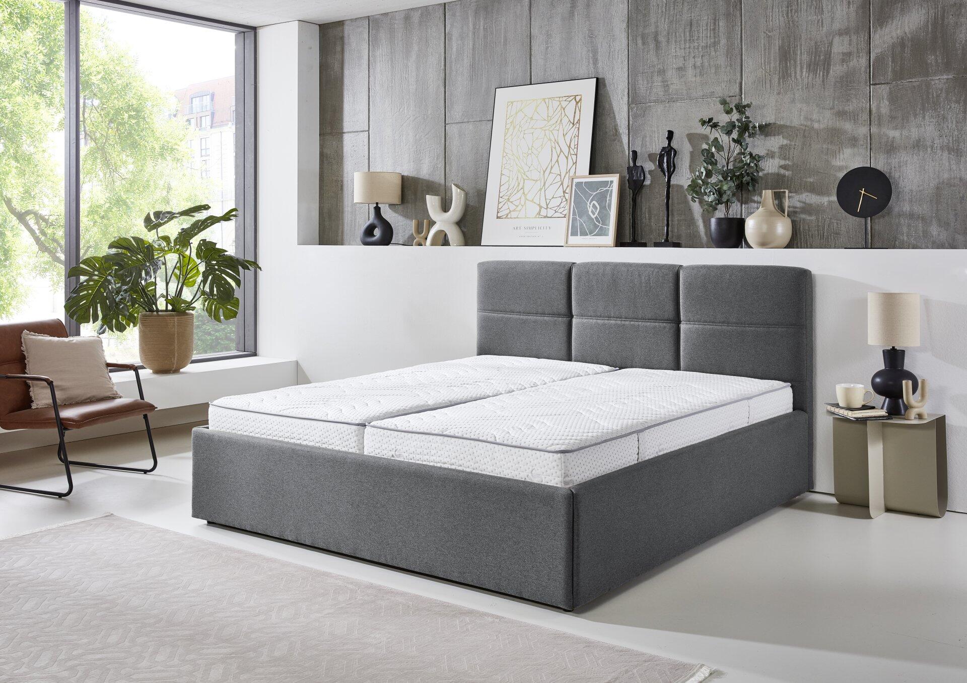 Polsterbett Durango PB100 mit grauem Stoffbezug, gepolstertem Kopfteil und sichtbaren Boxen unter der Matratze in einem stilvoll eingerichteten Schlafzimmer.