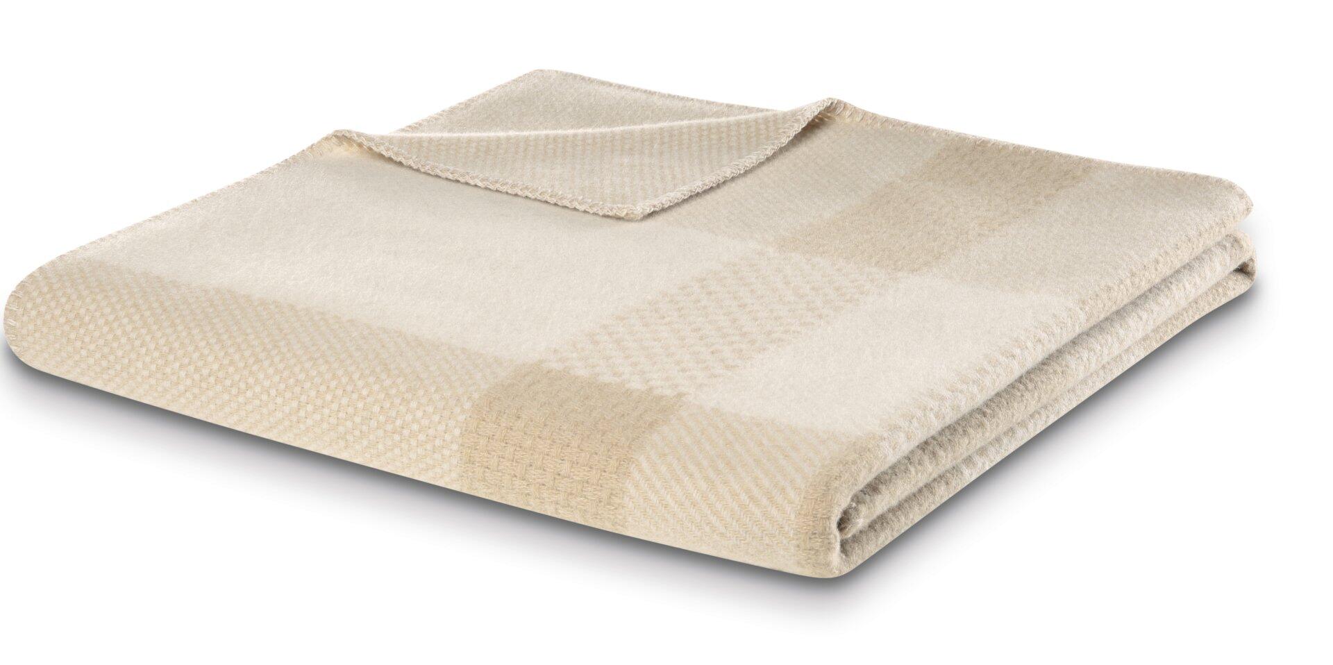 Plaid Crossweft beige: Detailaufnahme der gefalteten Tagesdecke aus 100% Wolle mit gewebter Textur und leicht schräg vor dem Hintergrund.