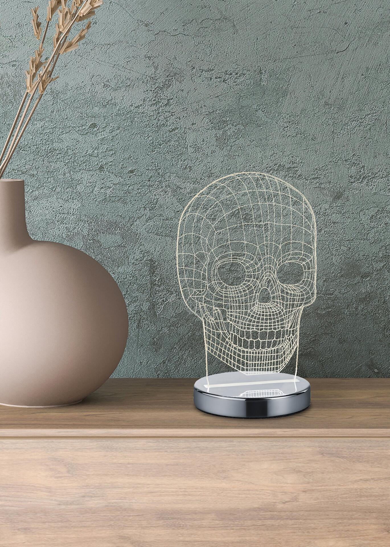 Dekorative Tischleuchte Skull mit silbernem Sockel und filigranem Metallschädel, dessen Struktur durch das Lichtmuster hervorgehoben wird.