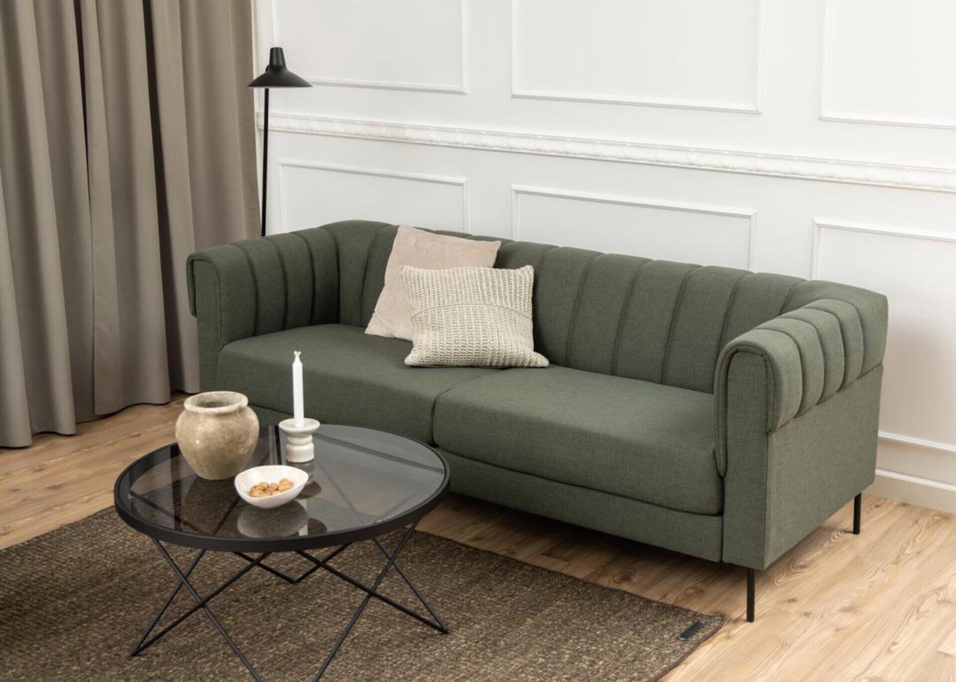 Couchtisch Cuxton mit schwarzem Glas und filigranem Metallgestell, stilvoll platziert vor einer dunkelgrünen, modernen Couch.