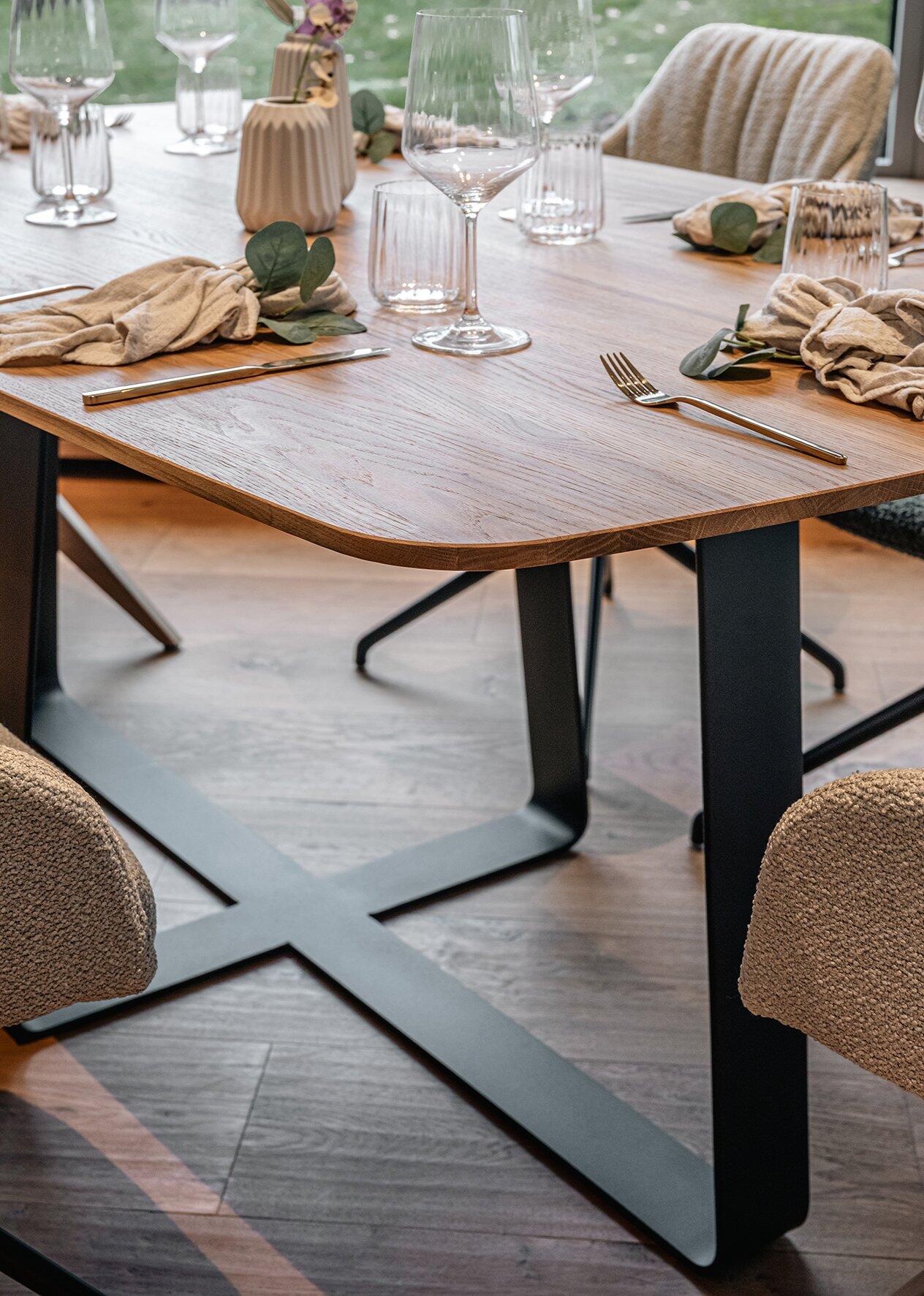 Esszimmertisch Urban mit bootsförmiger Eiche-Platte und schwarzem, X-förmigen Metallgestell, detailliert gezeigt mit gedecktem Tisch und stilvollen Accessoires aus leicht erhöhter Perspektive.