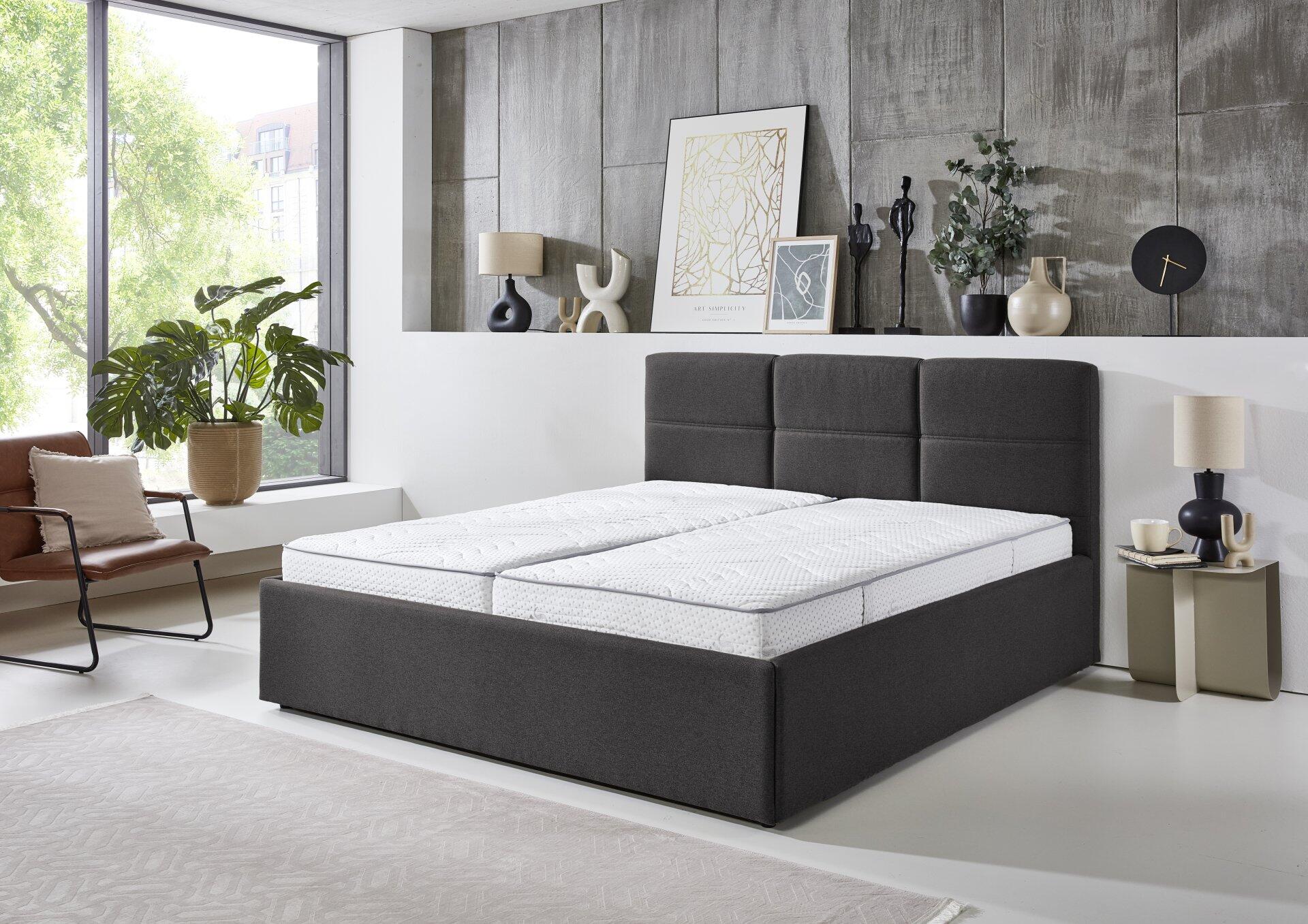 Graues Polsterbett Durango PB100 mit gestepptem Kopfteil und weißer Matratze in stilvollem Schlafzimmerambiente.