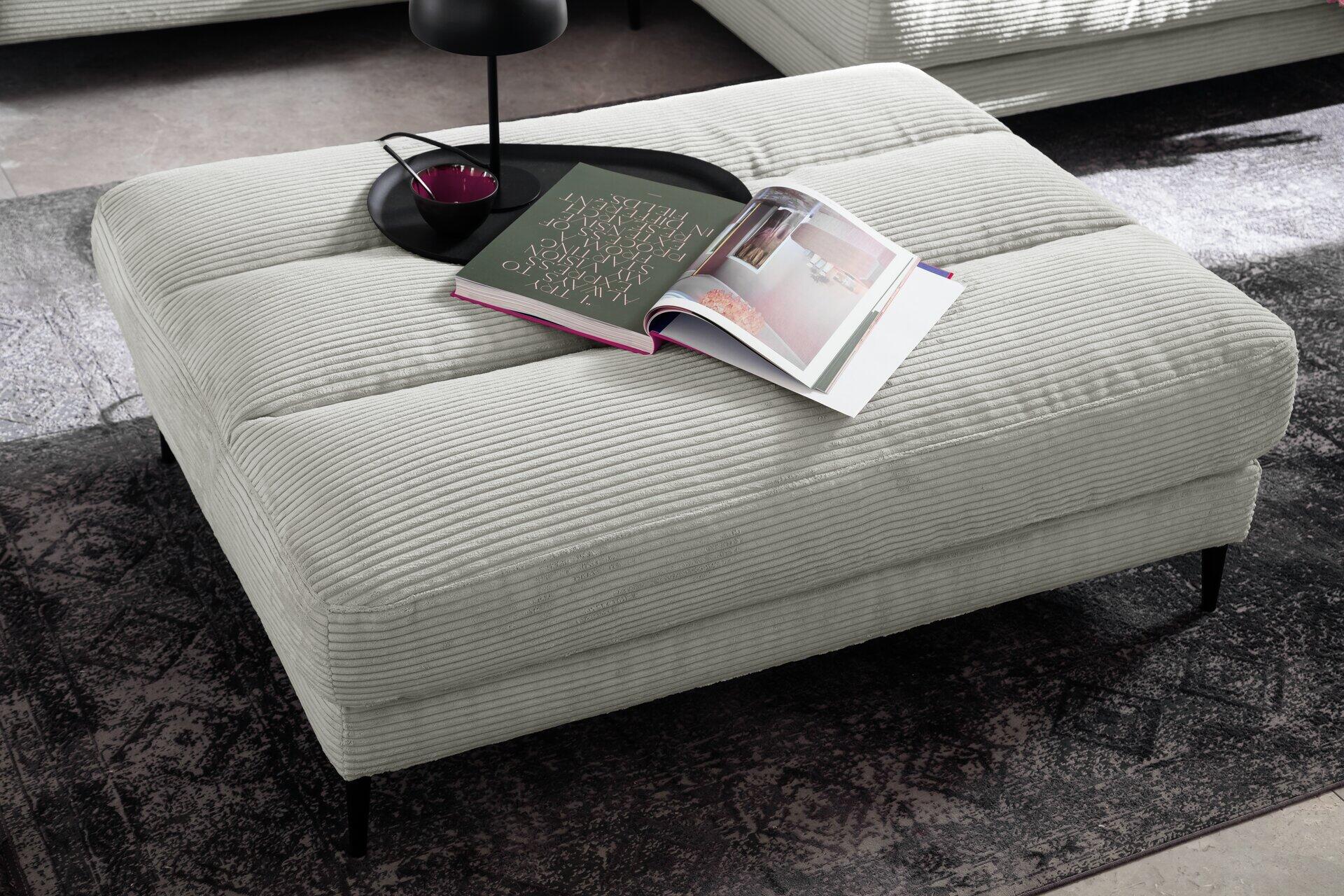 Hockerbank Badia mit grauem Cordstoffbezug und schwarzem Metallfuß, präsentiert als bequemer Sitzplatz und Ablagefläche mit offenem Buch und dekorativer Umgebung.