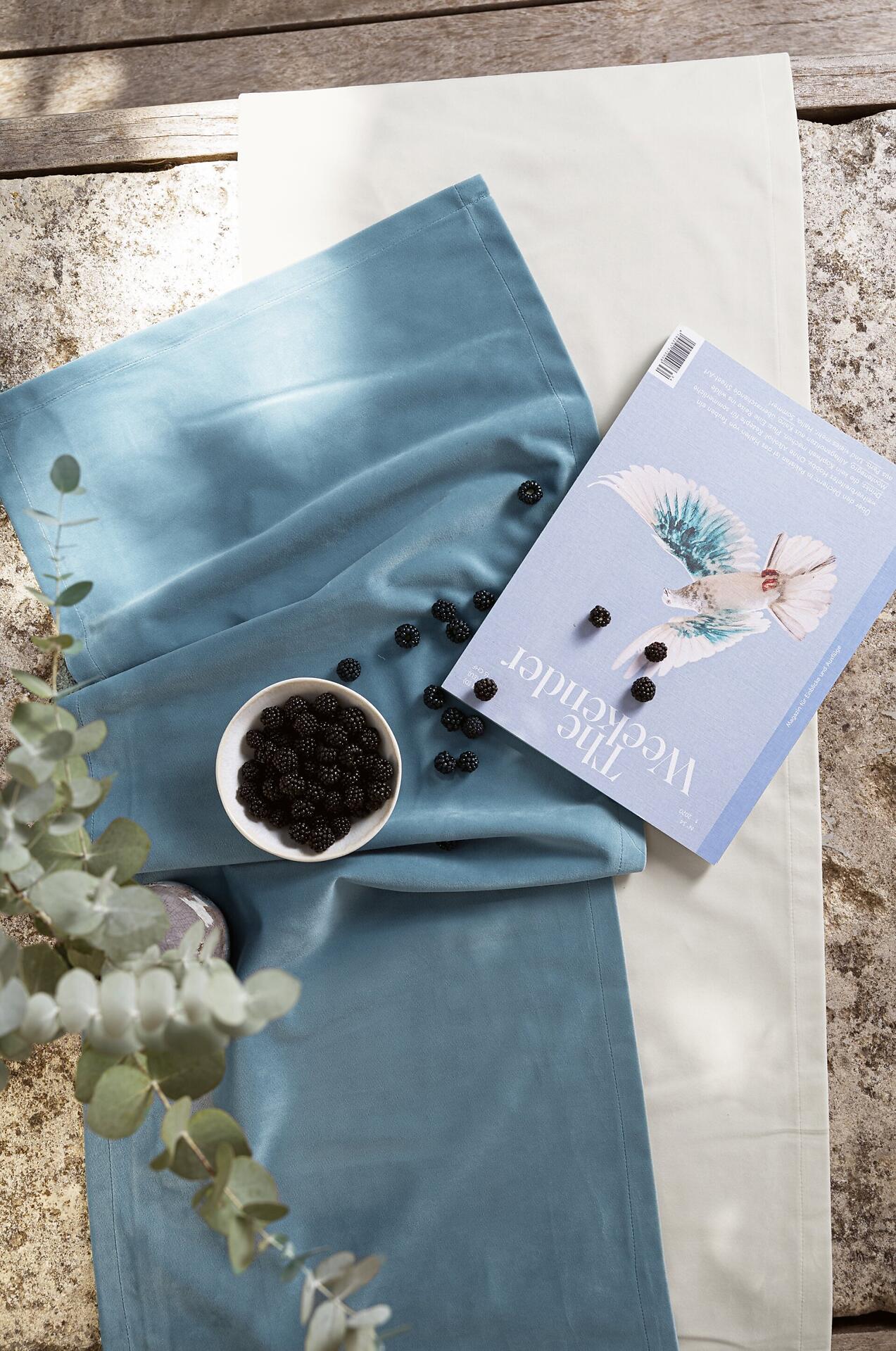 Blaugrau-farbener Samt-Tischläufer mit Brombeeren in einer Schale und Magazin, detailreich aus nahaufnahme Perspektive fotografiert.