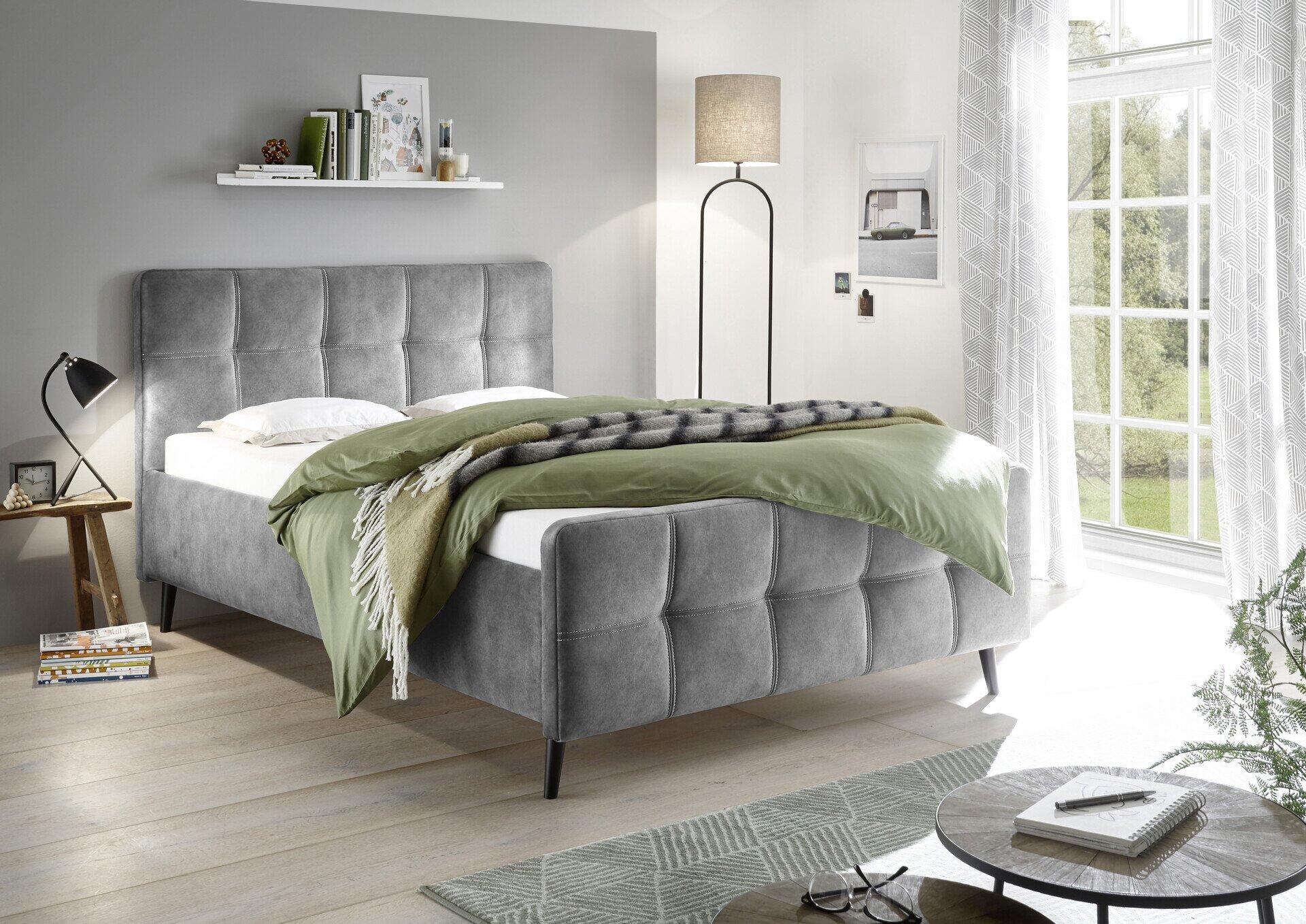 Modernes Polsterbett Mario C mit grauem Samtbezug, geometrischem Kopfteil und schlanken schwarzen Metallfüßen, abgebildet in einer Frontalansicht im hellen Schlafzimmer.