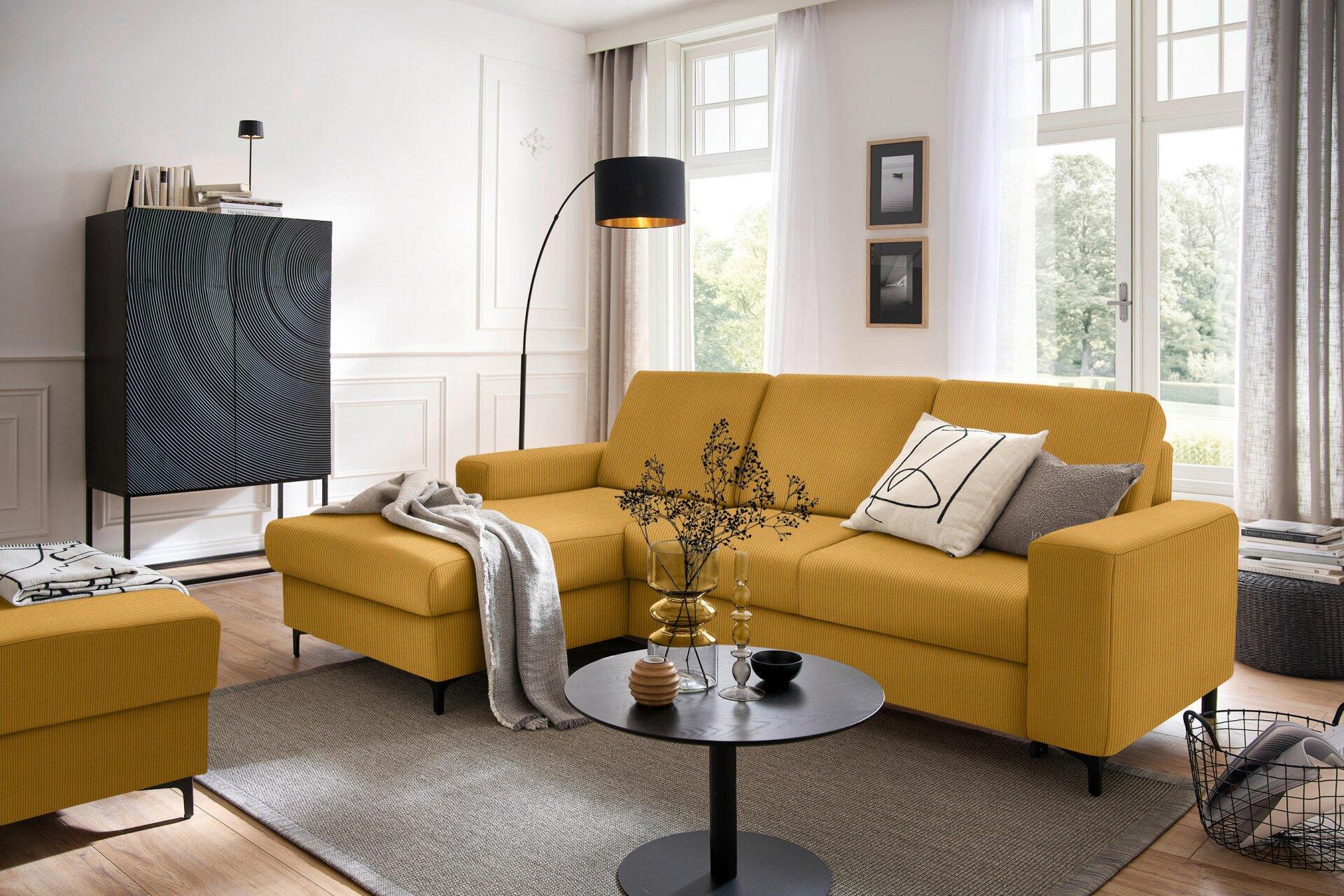 Ecksofa Balina in gelb-orangefarbener Stoffausführung mit Winkelfüßen, Bettkasten und Ottomane, dargestellt in einer stilvoll eingerichteten Wohnszene mit Fußhocker und passendem Stricküberwurf.