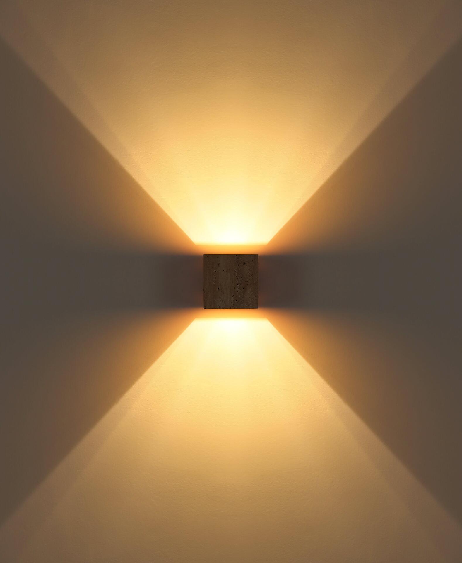 Wandleuchte ROJAS mit Travertin-Glas, die in einer Nahaufnahme die flach an der Wand montierte, quadratische Form mit rustikaler Holzoptik und die nach oben und unten abstrahlenden Lichtstrahlen zeigt, die geometrische Lichtlinien auf der Wand erzeugen.