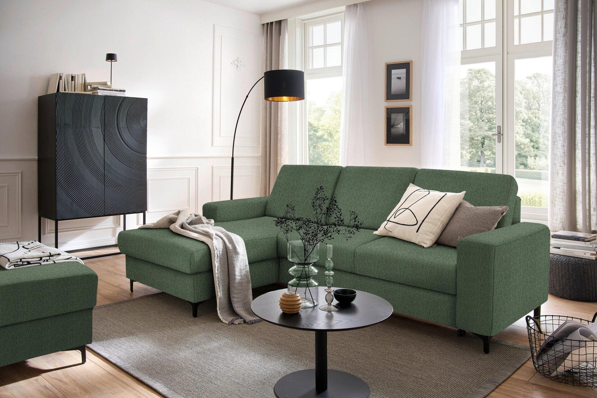 Ecksofa Balina in dunkelgrün mit Winkelfüßen und schlichtem Design, präsentiert in einem hellen Wohnzimmer mit rundem Couchtisch und dekorativen Elementen.