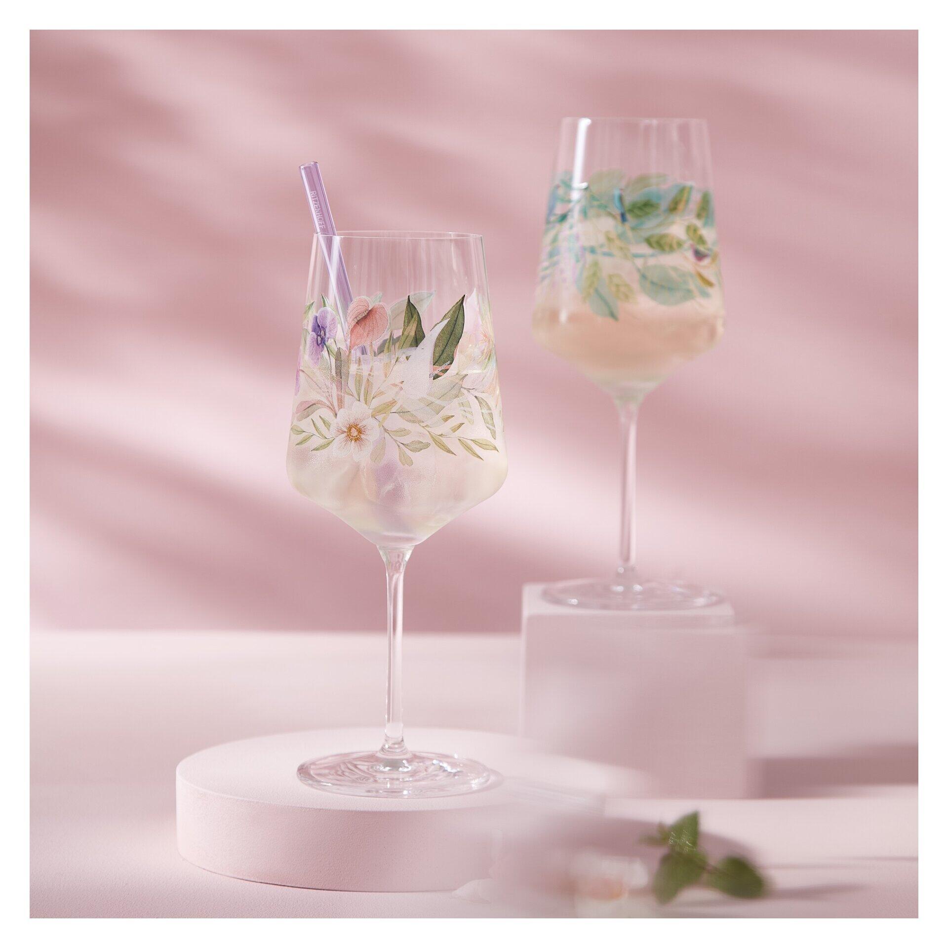 Zwei hoch-schlanke Aperitifgläser mit floralem Dekor und einem rosafarbenen Getränk, präsentiert auf einem rosafarbenen Podest.