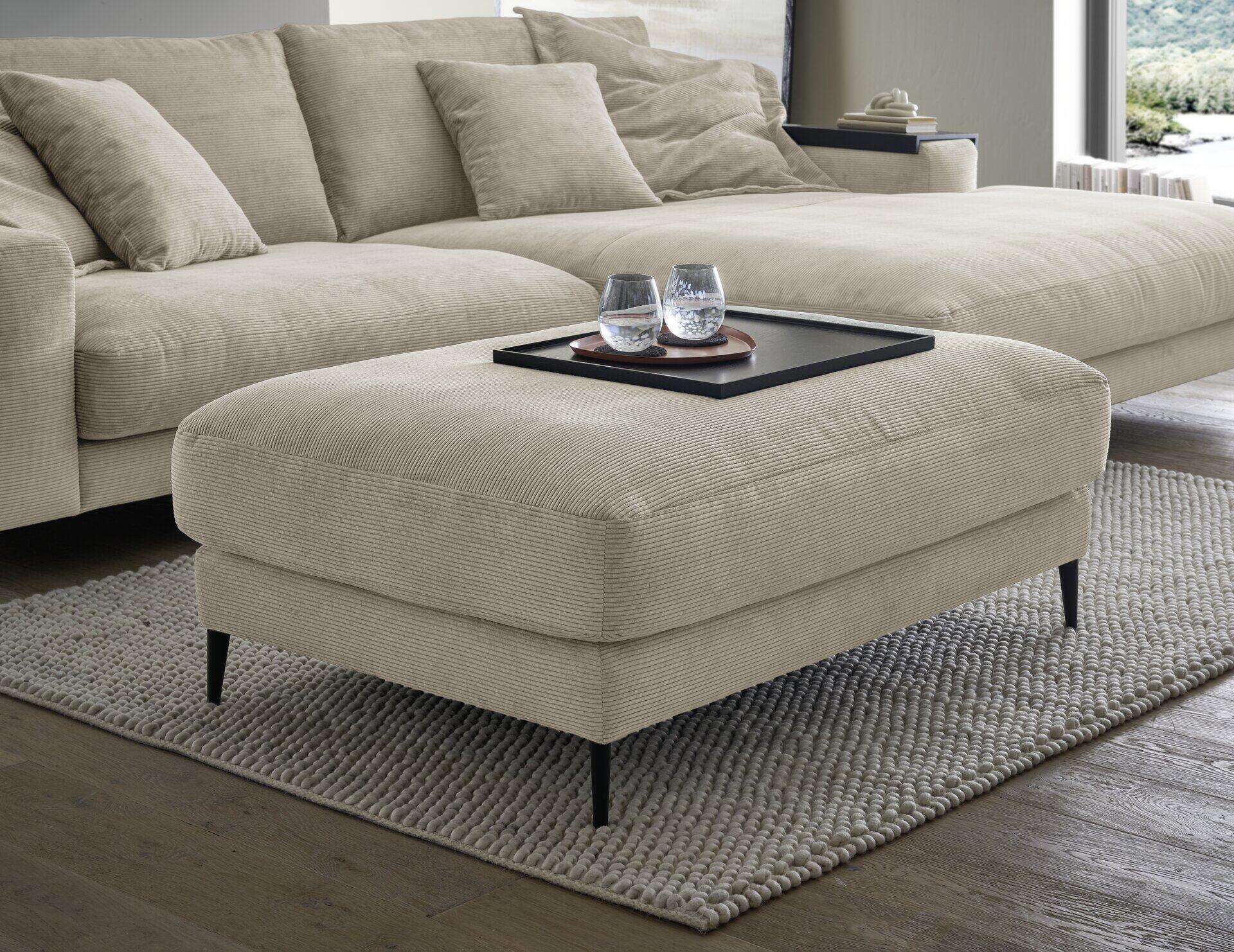 Fußhocker Badia Base 2.0 mit gerippter Oberfläche in Hellbeige auf schlanken schwarzen Metallbeinen, präsentiert vor einer Couch und Teppich, aus leicht erhöhter Perspektive.