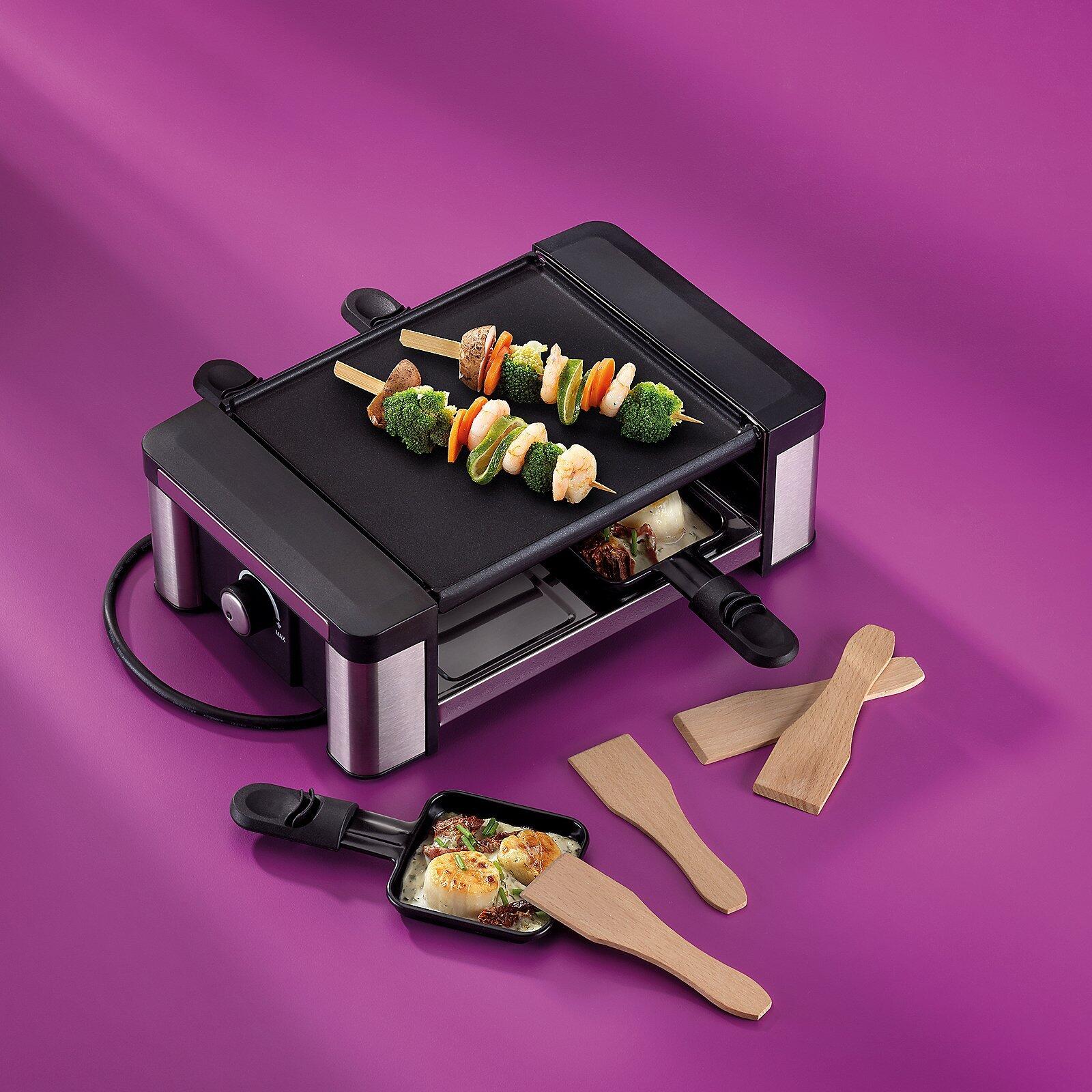 Raclette/Grillplatte Timur mit silbernem Gehäuse, schwarzer Antihaftbeschichtung und zwei vertikal gegenüberliegenden Grillbereichen, auf denen Fleisch- und Gemüsespieße liegen.