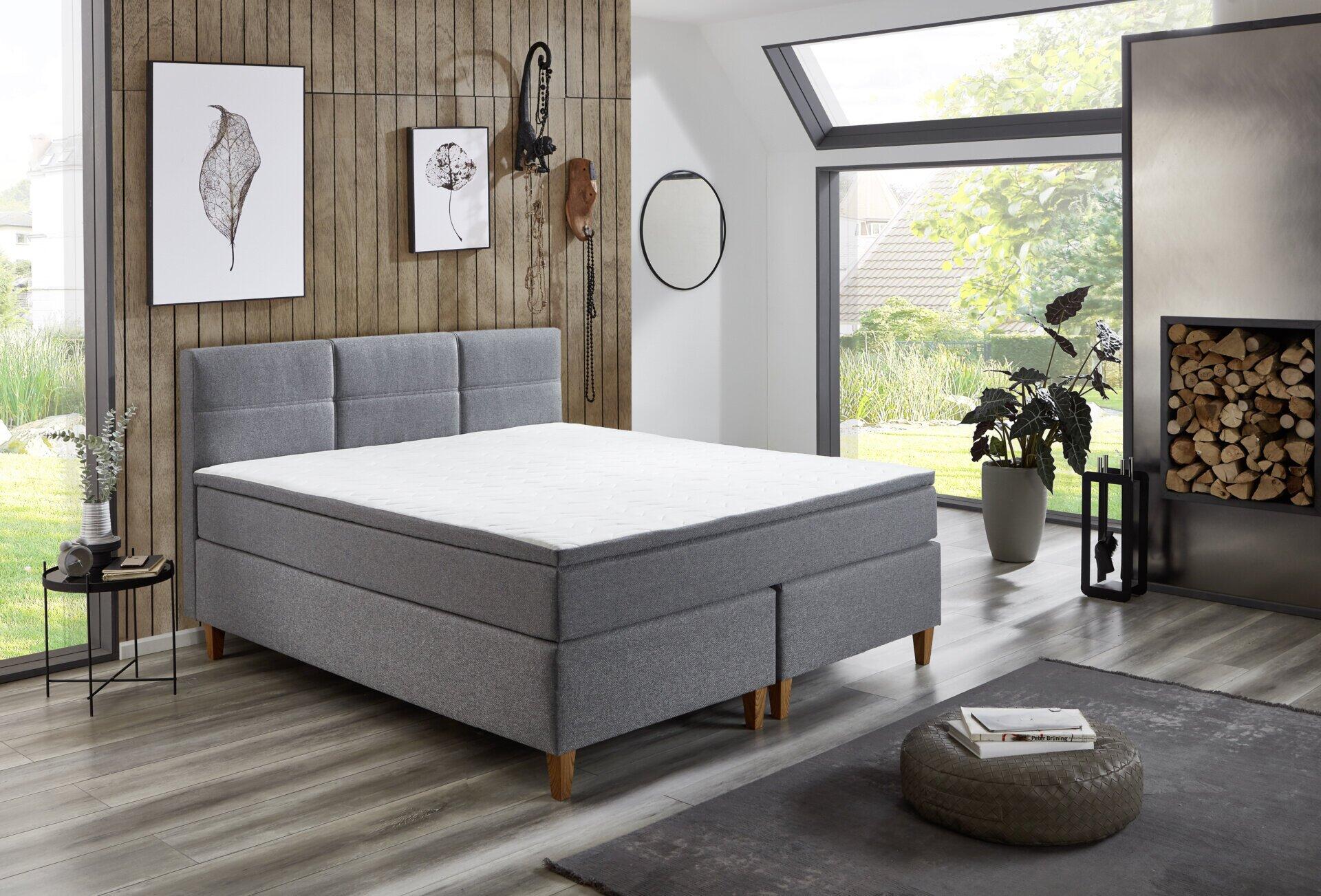 Boxspringbett Kenora BX2360 in Seitenansicht mit grauem Stoffbezug, Holzfüßen und Stauraum durch erhöhte Matratzenposition.