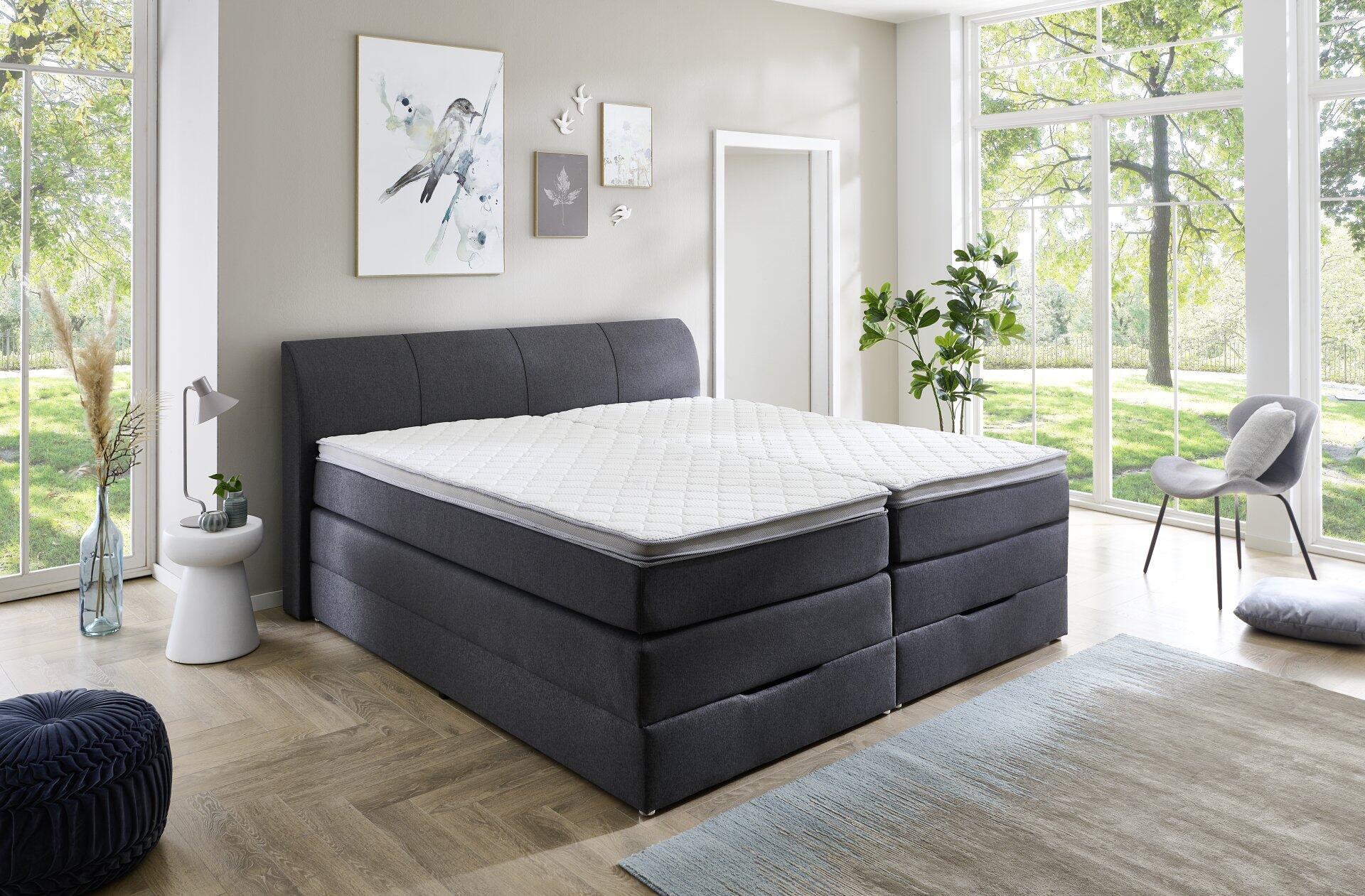 Graues Boxspringbett Modesto BX1440 mit integrierten Schubladen für Stauraum und hellem, gesteppten Bezug, frontal abgebildet in einer hellen Schlafzimmereinrichtung.