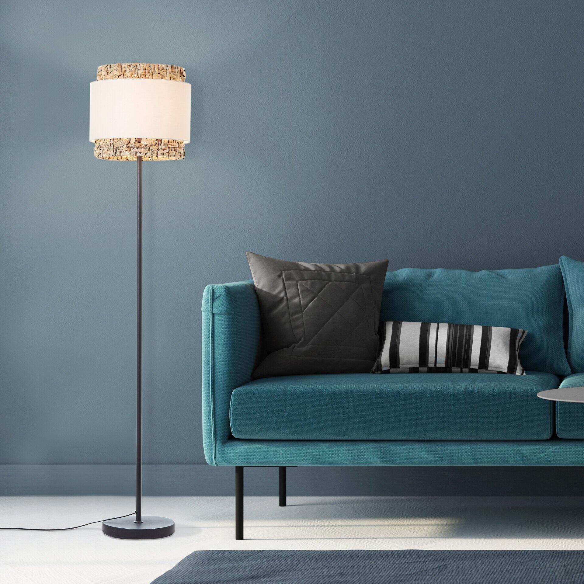 Stehleuchte Waterlilly mit schwarzem Metallfuß und zylindrischem Lampenschirm aus Rattan neben einem blauen Sofa.