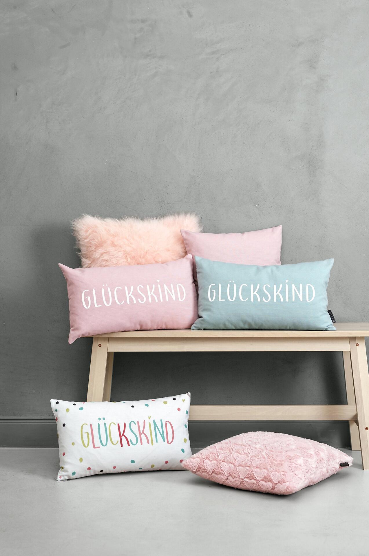 Drei Glückskind Fino Kissen mit unterschiedlichen Designs und dem Schriftzug "GLÜCKSKIND" auf einem hellen Holzstuhl arrangiert.